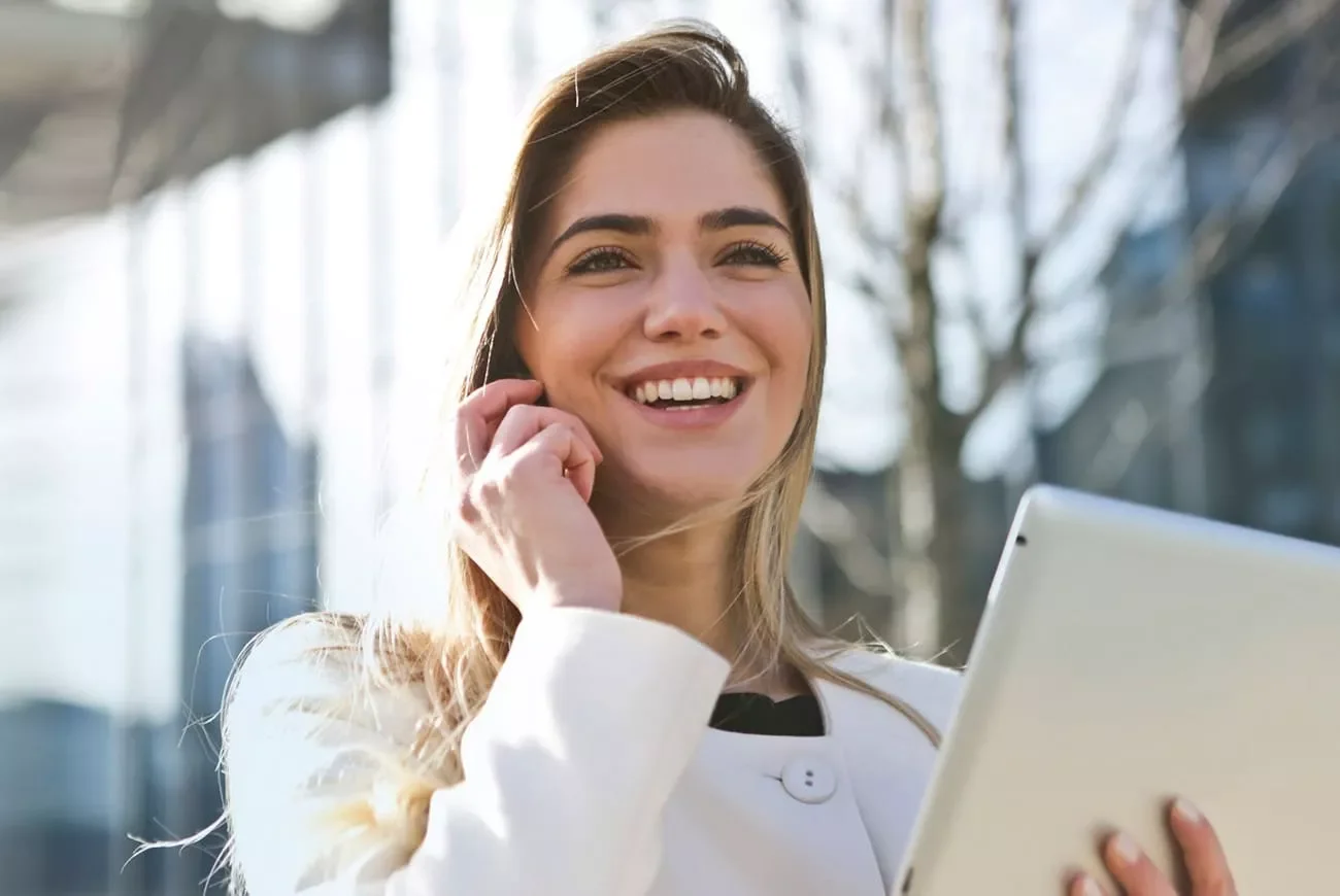 A smiling young woman holding a tablet outdoors with trees and a modern building in the background.