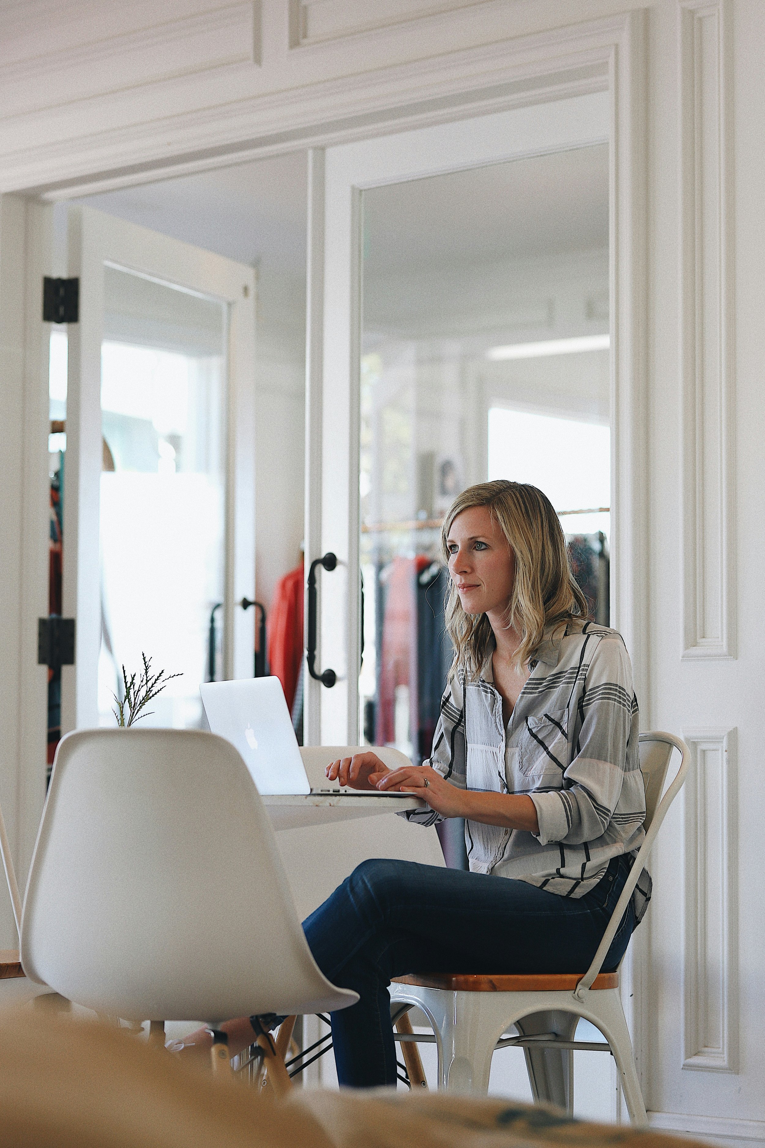 A woman with blonde hair working on a laptop at a dining table in a bright room with white walls and chairs, and a window in the background.