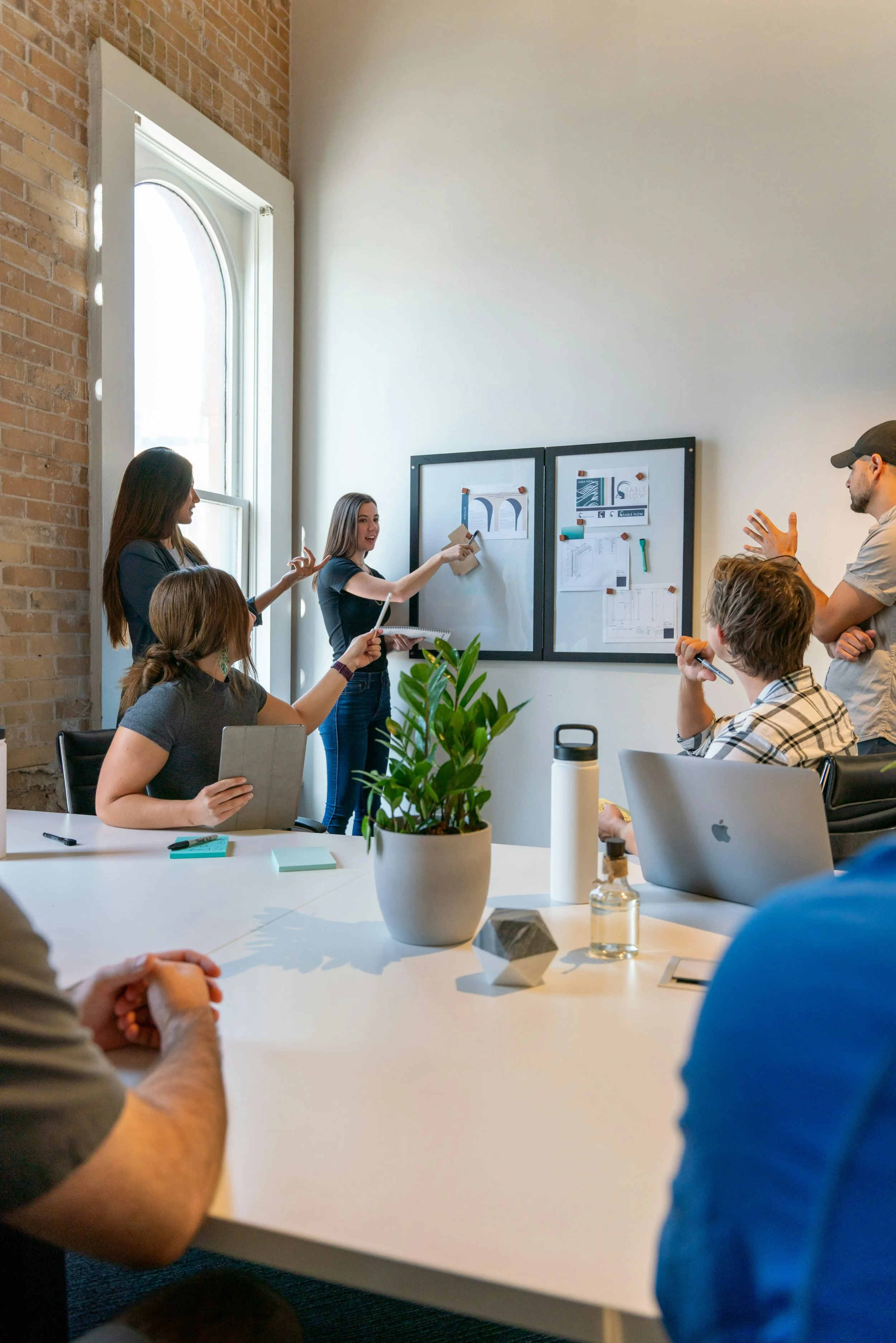 A group of people in a meeting room with exposed brick walls and white walls. One woman is standing and presenting, pointing at a whiteboard with diagrams and papers, while others are sitting around a white table with laptops, notebooks, and a potted plant.