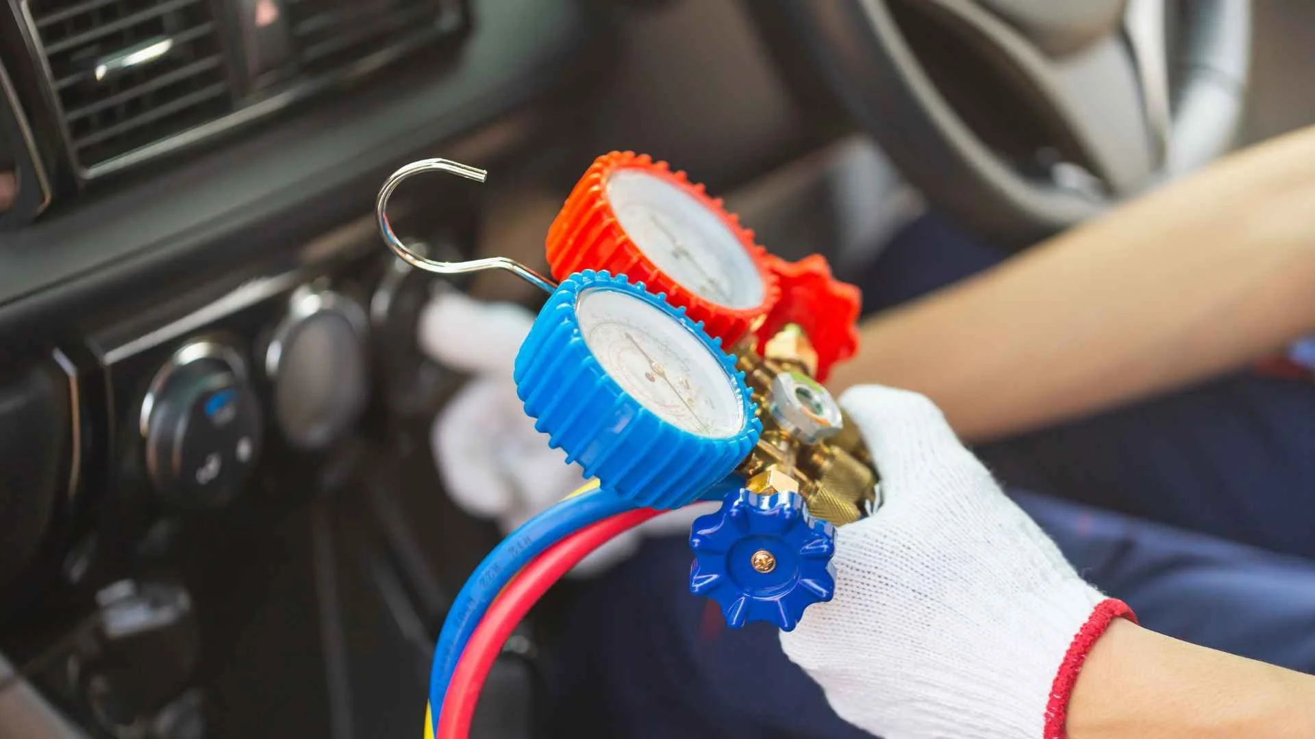 A person wearing a white glove is holding a refrigerant manifold gauge set inside a vehicle with the dashboard and controls visible in the background.