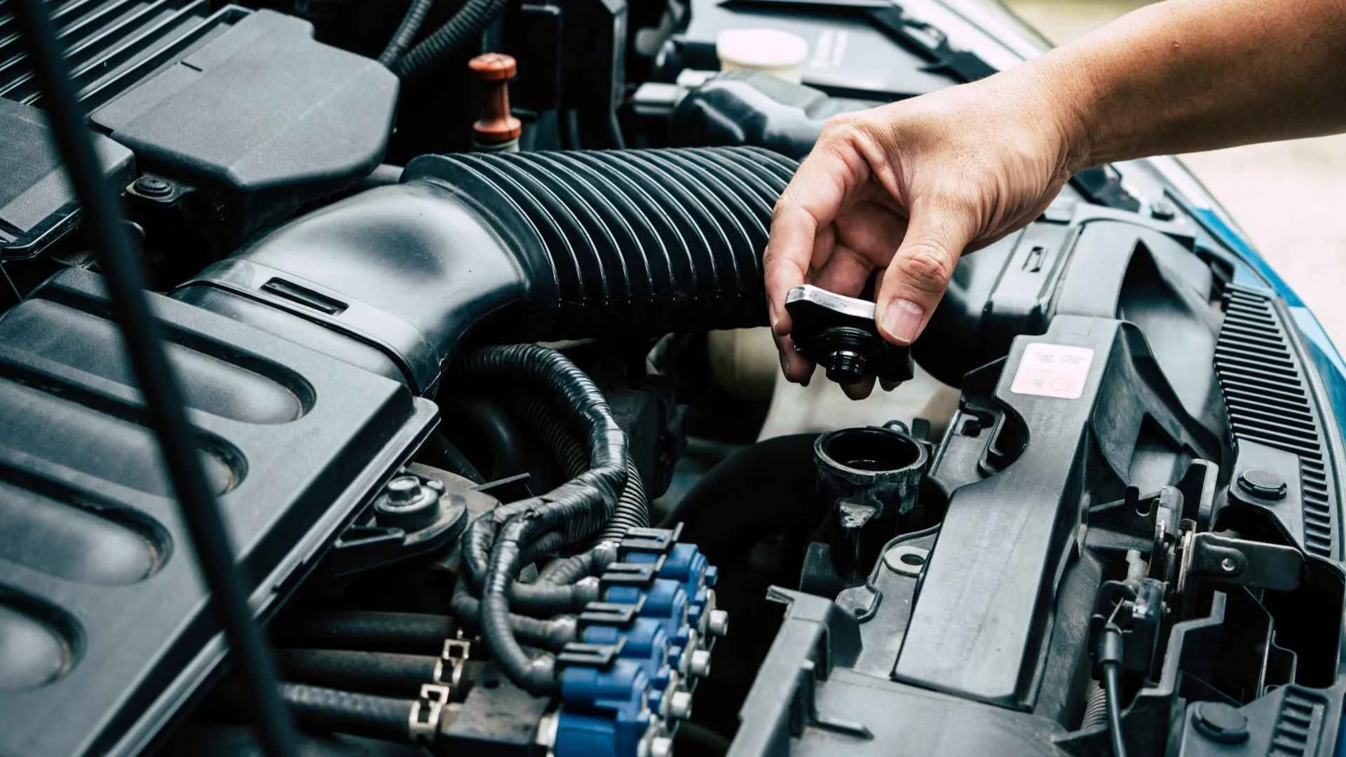 Close-up of a person's hand removing a cap from an engine fluid reservoir under the hood of a car, with various engine parts visible.
