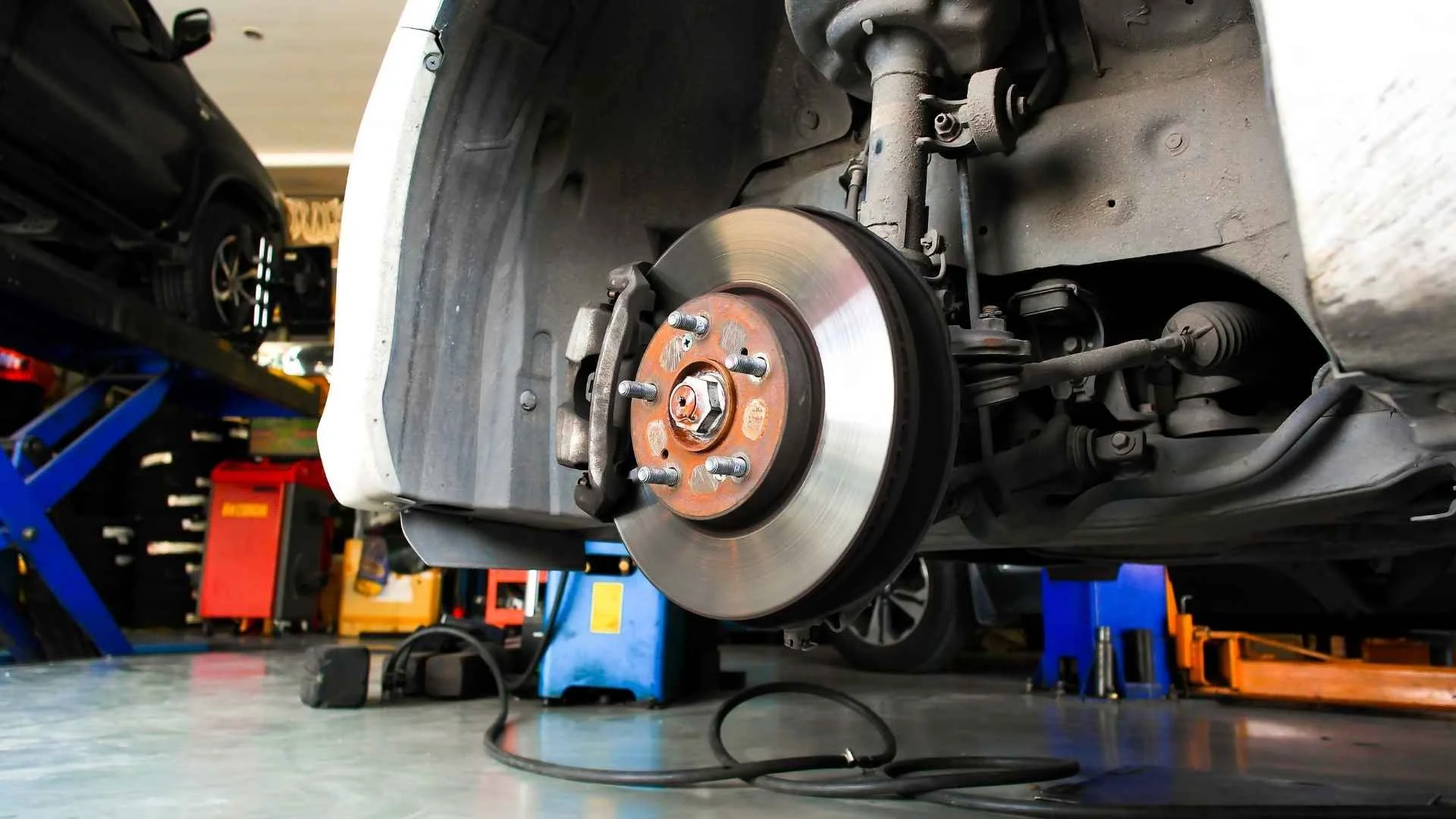 Close-up of a car’s front brake rotor and suspension on a lift in an auto repair shop, showing a winter brake inspection in progress.