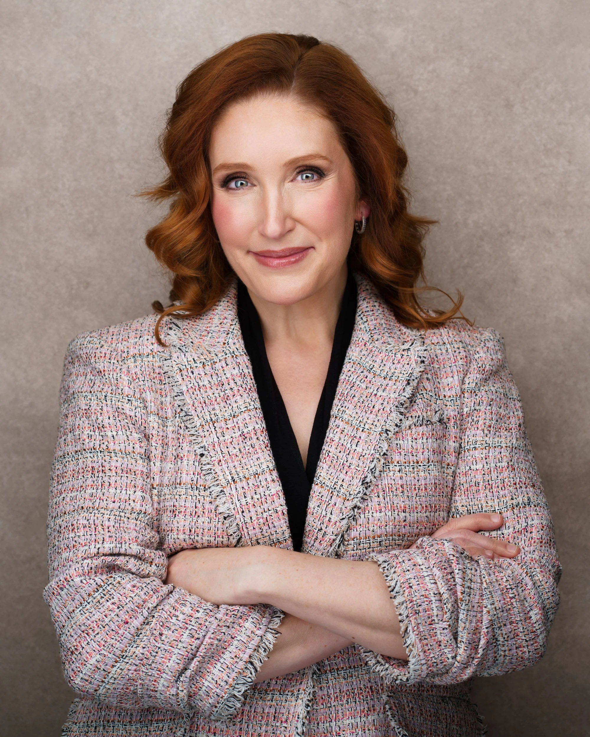 Tissa Richards wearing a pink and white tweed blazer, standing against a plain beige background, with her arms crossed and a confident smile.