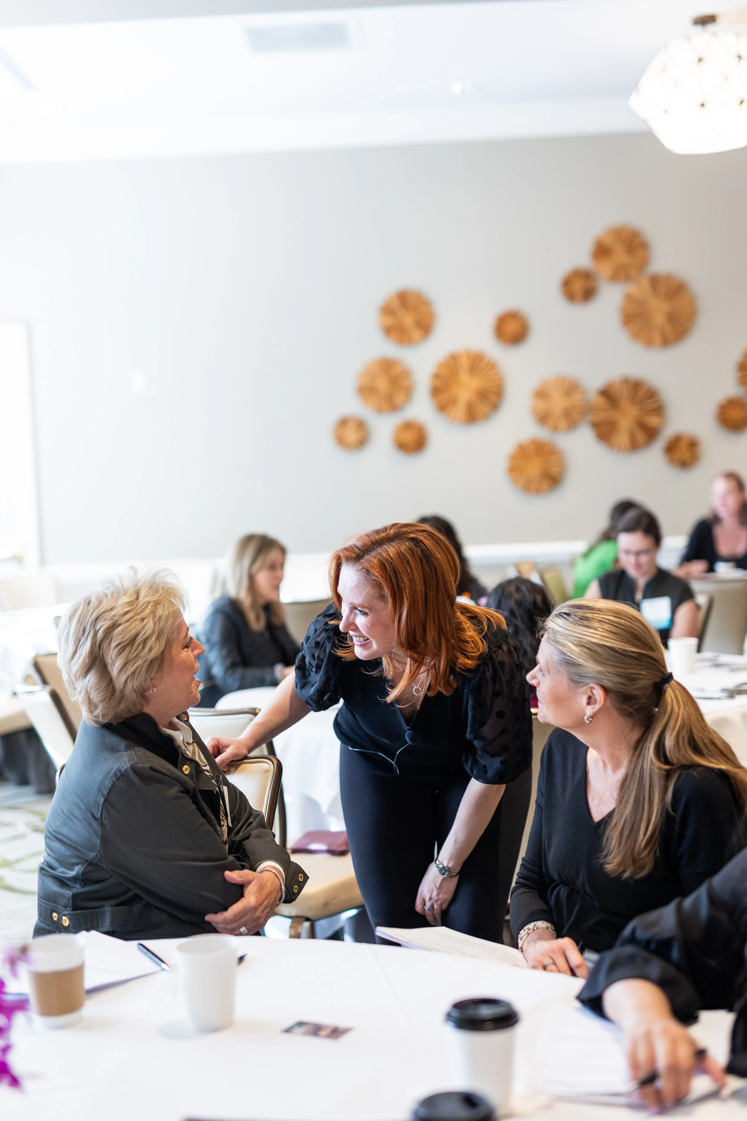 Tissa Richards smiling and talking to an older woman with blonde hair during a meeting in a conference room with other attendees in the background.