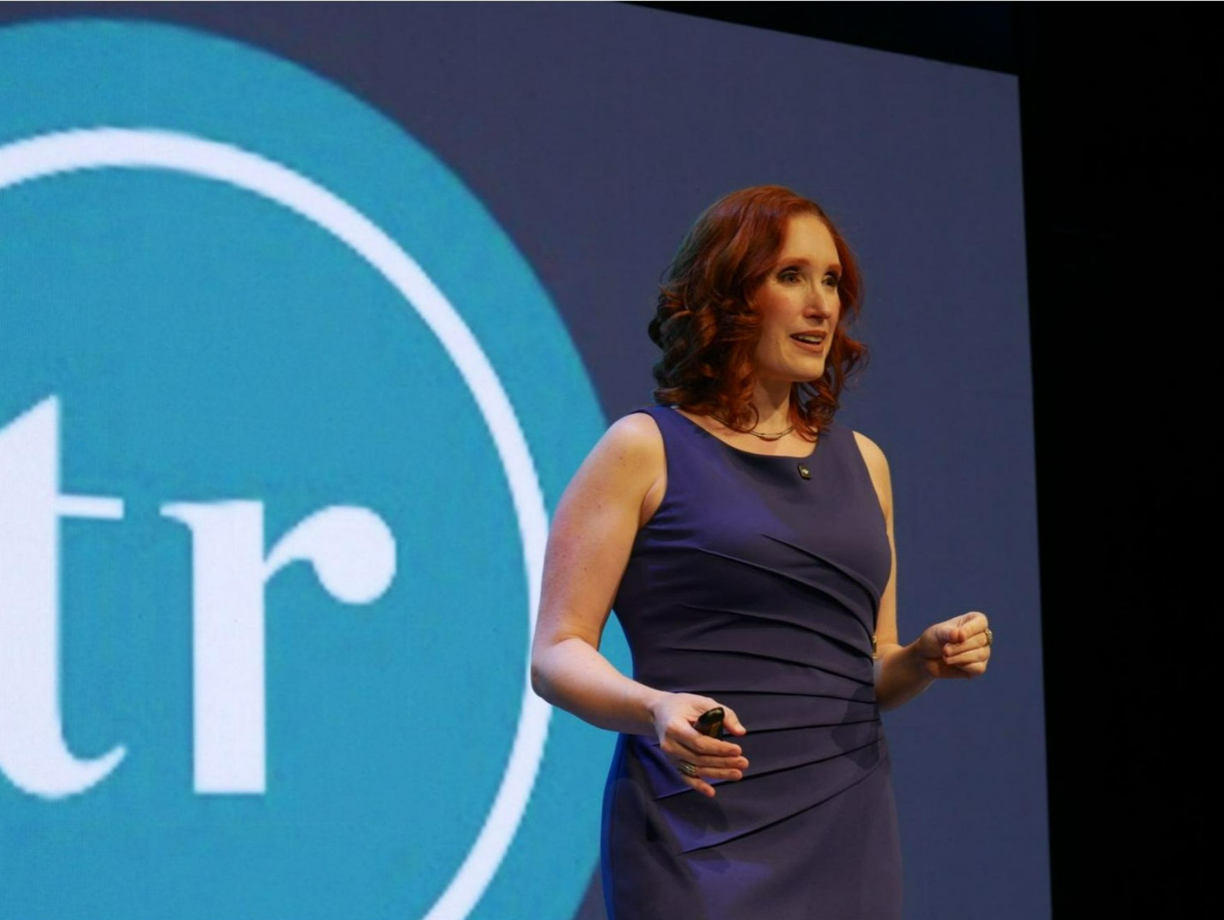 Tissa Richards wearing a navy blue sleeveless dress, speaking on stage in front of a large presentation screen.