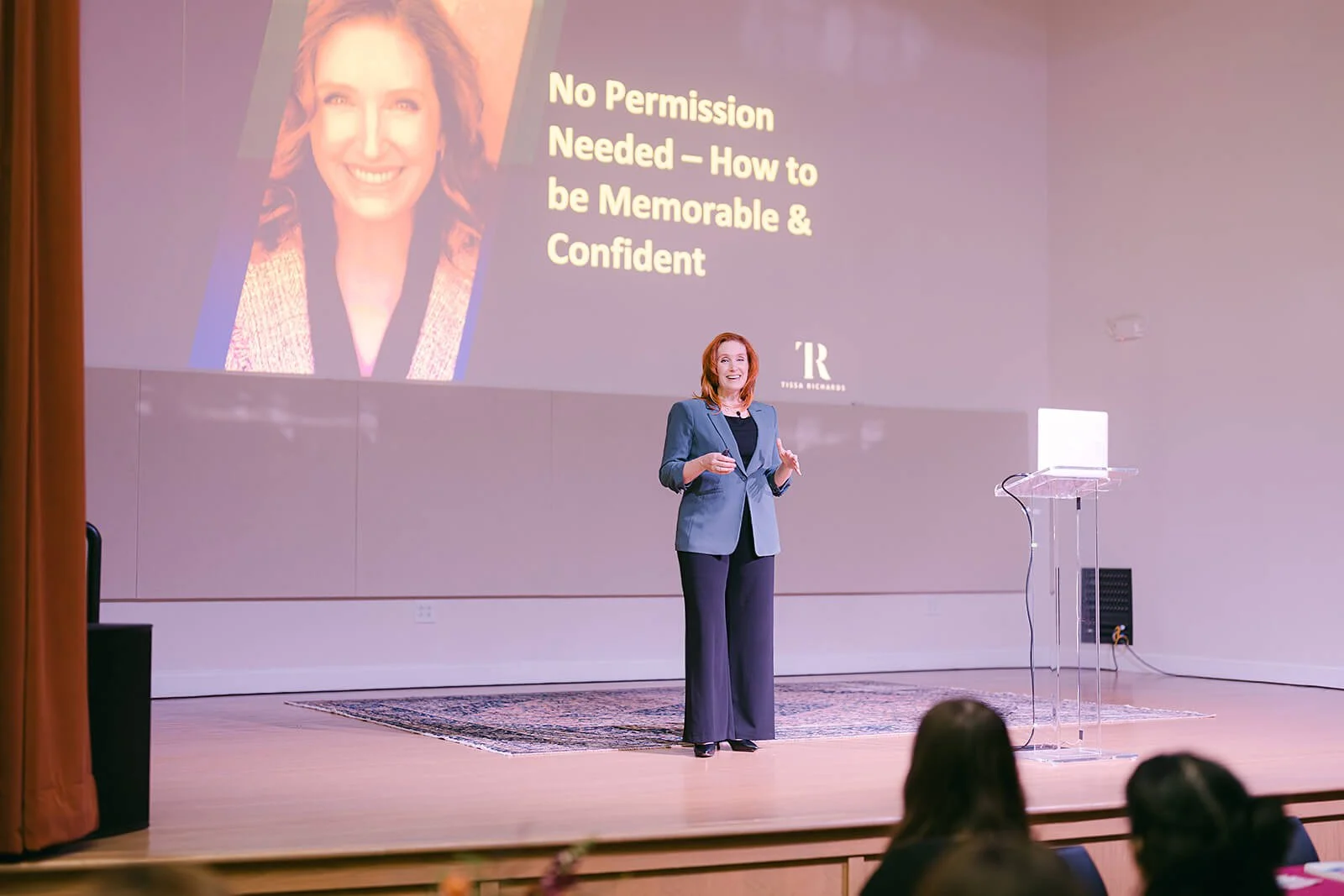 Tissa Richards stands on a stage giving a presentation with a large screen behind her displaying the title 'No Permission Needed — How to be Memorable & Confident' and a photo of her smiling.