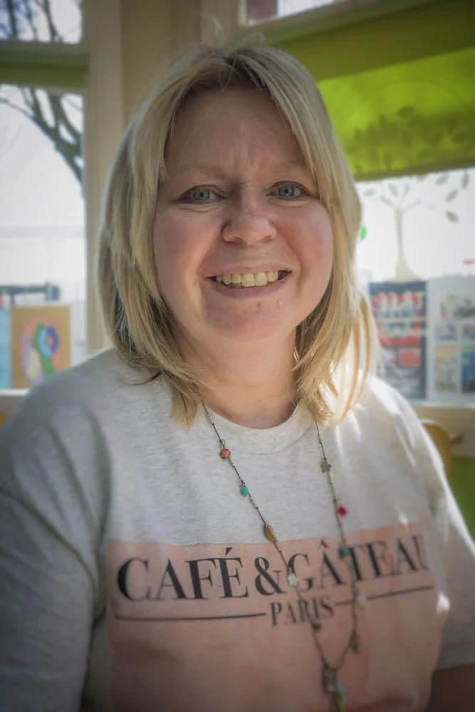 A woman with blonde hair smiling, wearing a light gray T-shirt with 'Café & Gâteau Paris' printed on it, and a beaded necklace, inside a brightly lit cafe or shop with display items and artwork in the background.