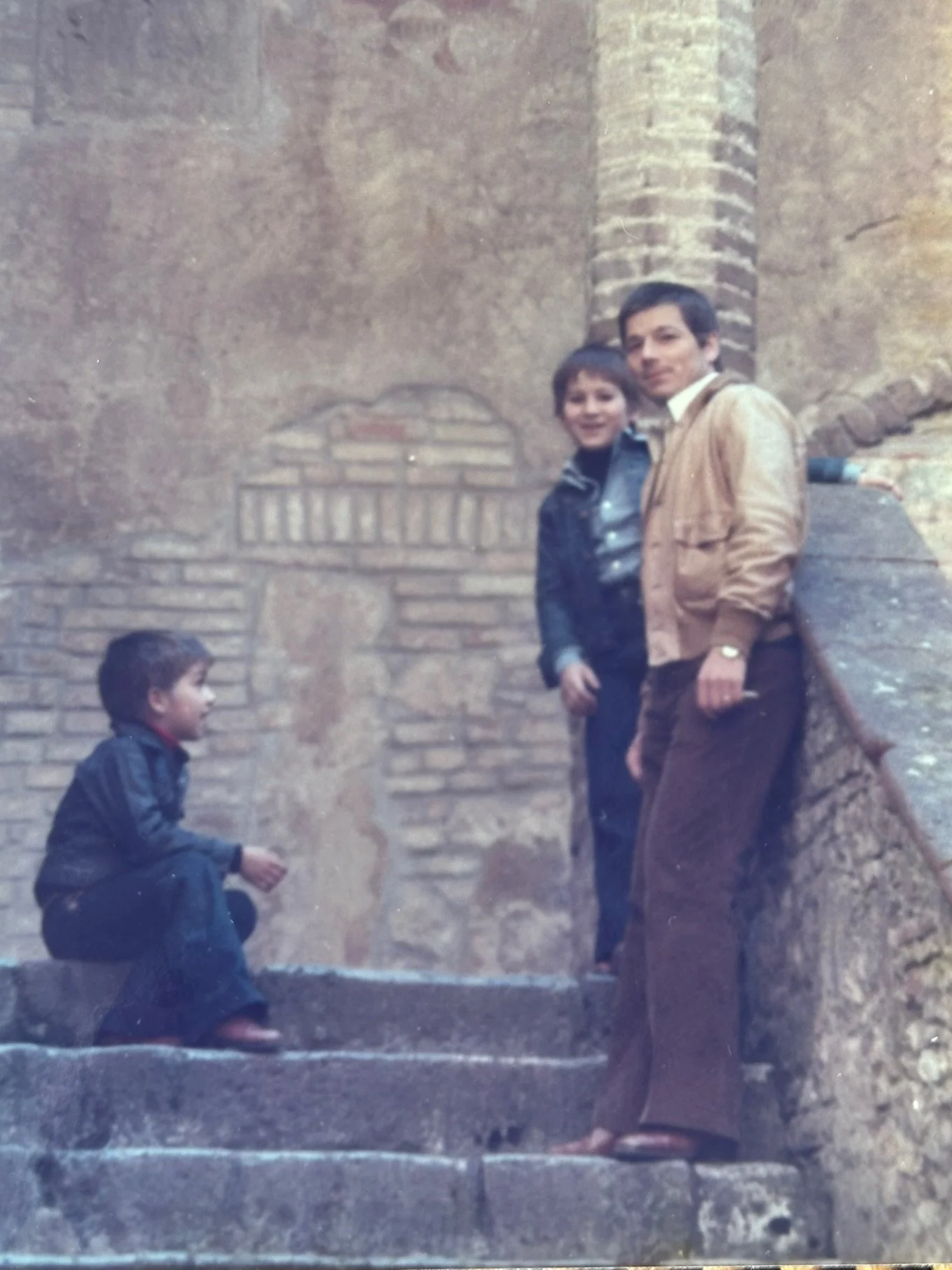 Three boys on stone steps outside a brick and stone wall, two standing and one crouching.