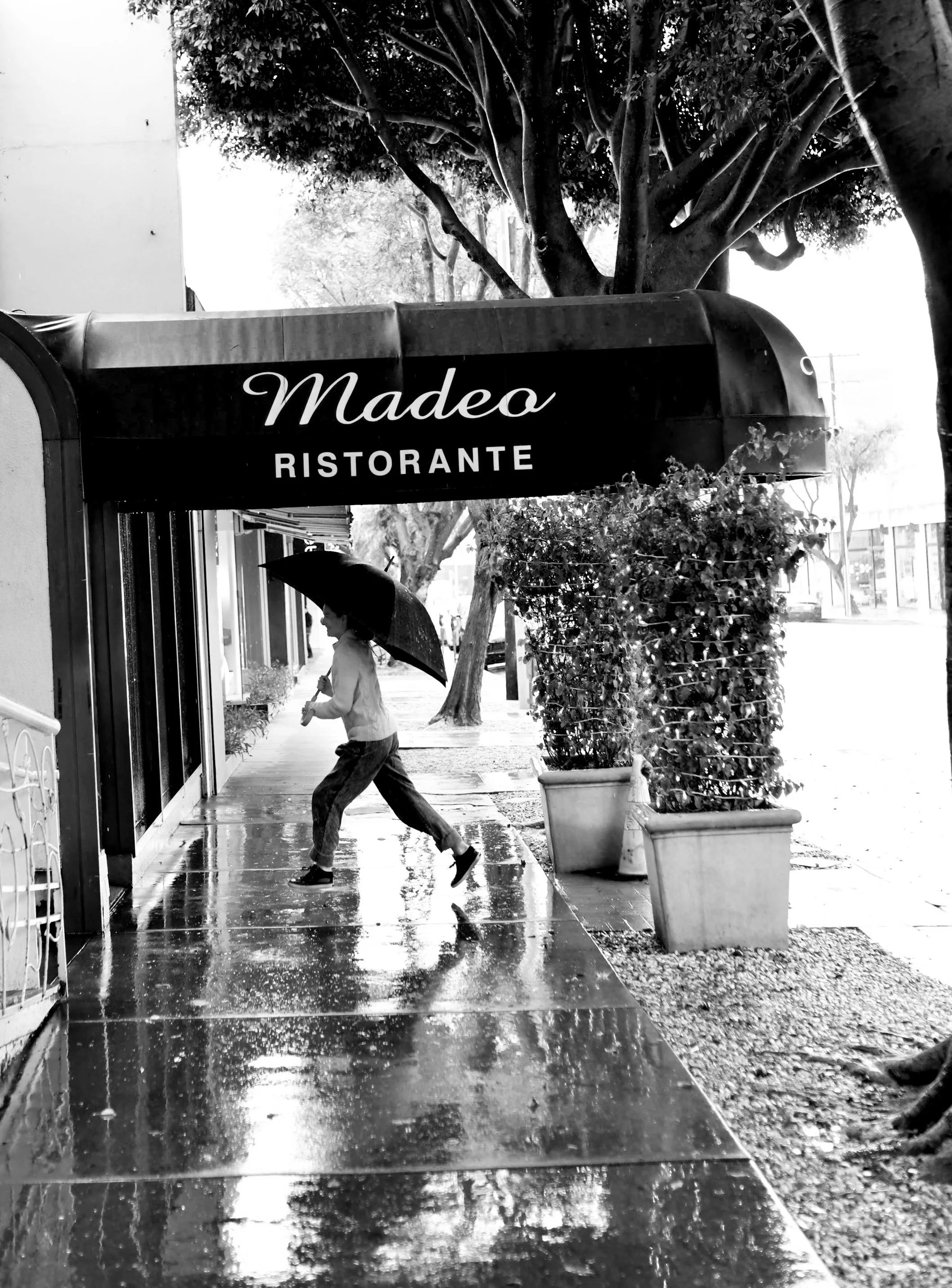 A person running on a rainy sidewalk holding an umbrella outside a restaurant called Madeso Ristorante, with trees and potted plants along the street.