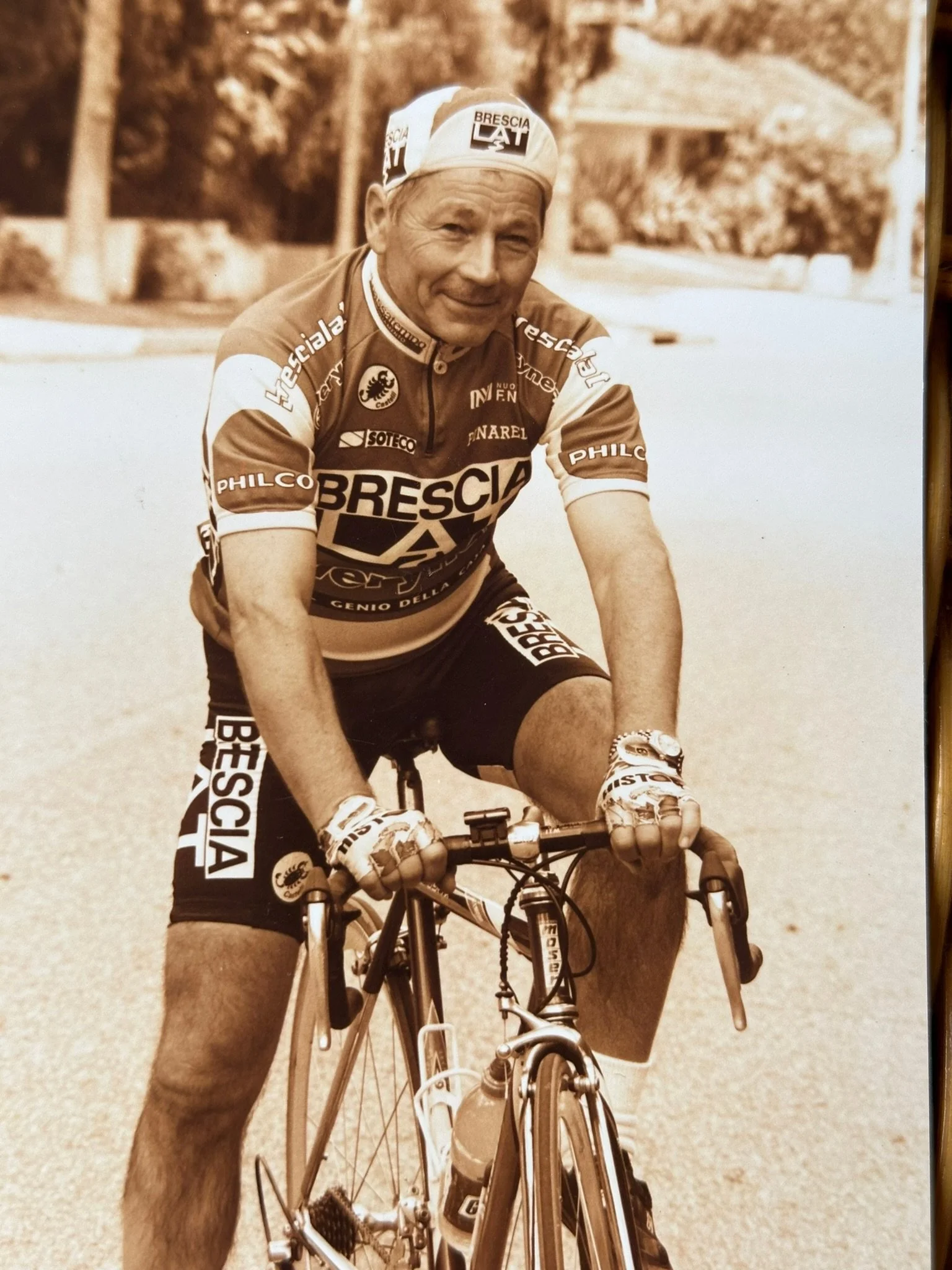 An elderly man wearing a Brescia cycling team jersey and cap, riding a bicycle outdoors on a paved surface.