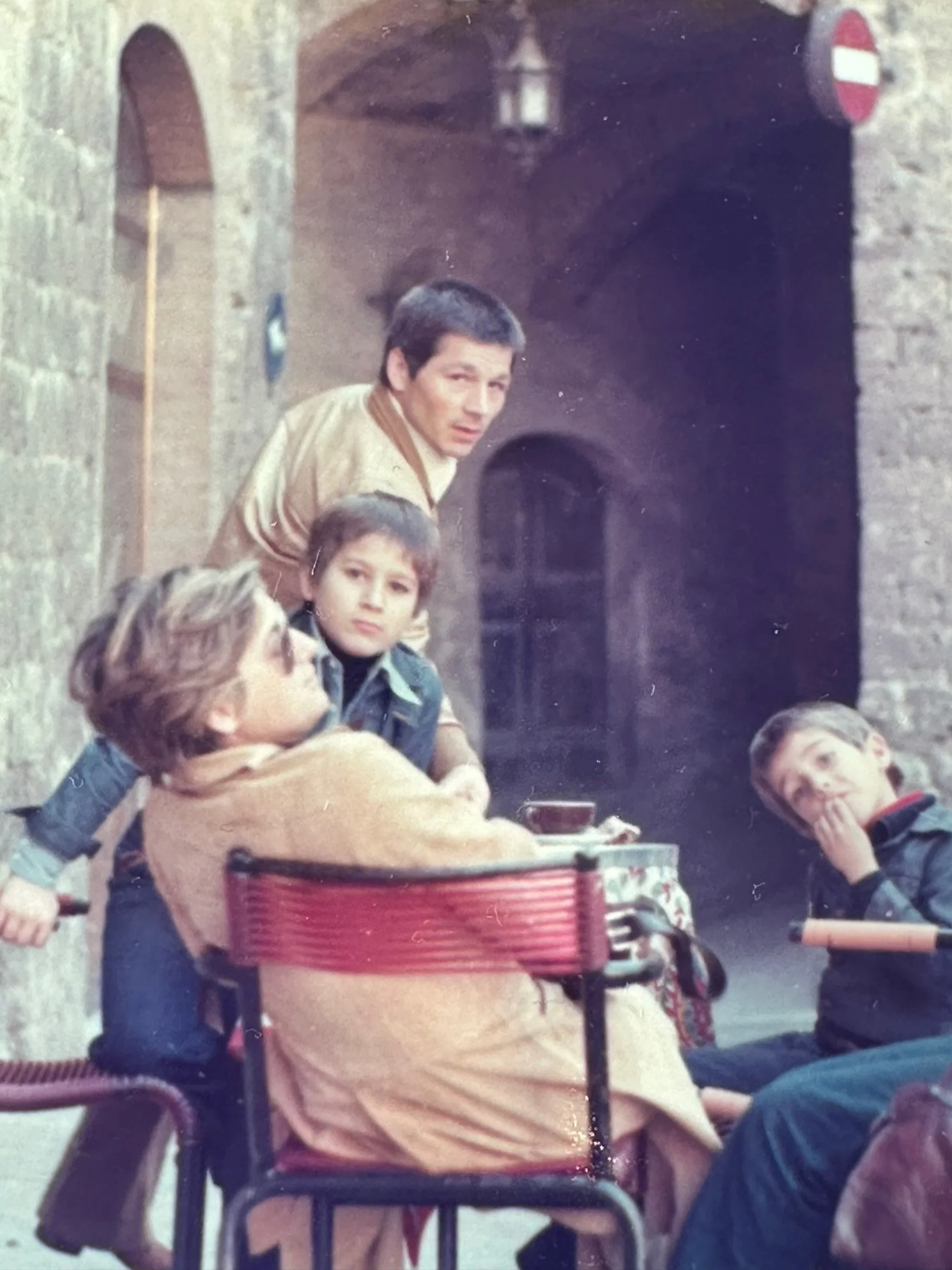 Four children sitting outdoors in front of a brick building; one girl in a wheelchair, two boys standing, and one boy sitting. The girl in the wheelchair is wearing glasses and a beige coat. The boy standing behind her has short dark hair, and the boy beside him has wet hair. The boy sitting has his hand over his mouth. The scene appears to be from a past decade, possibly the 1980s.