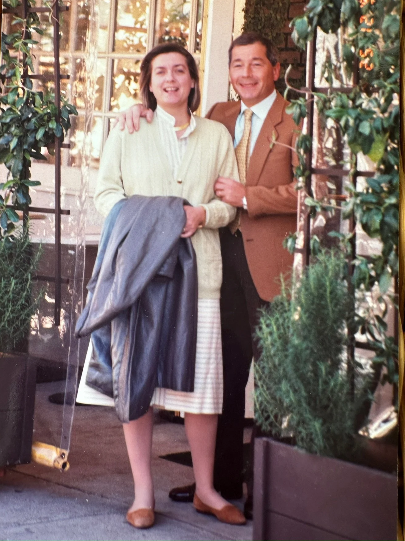 A woman and a man standing outdoors, smiling, with the woman holding a coat. They are framed by greenery and a brick column, and appear to be on a patio or terrace.