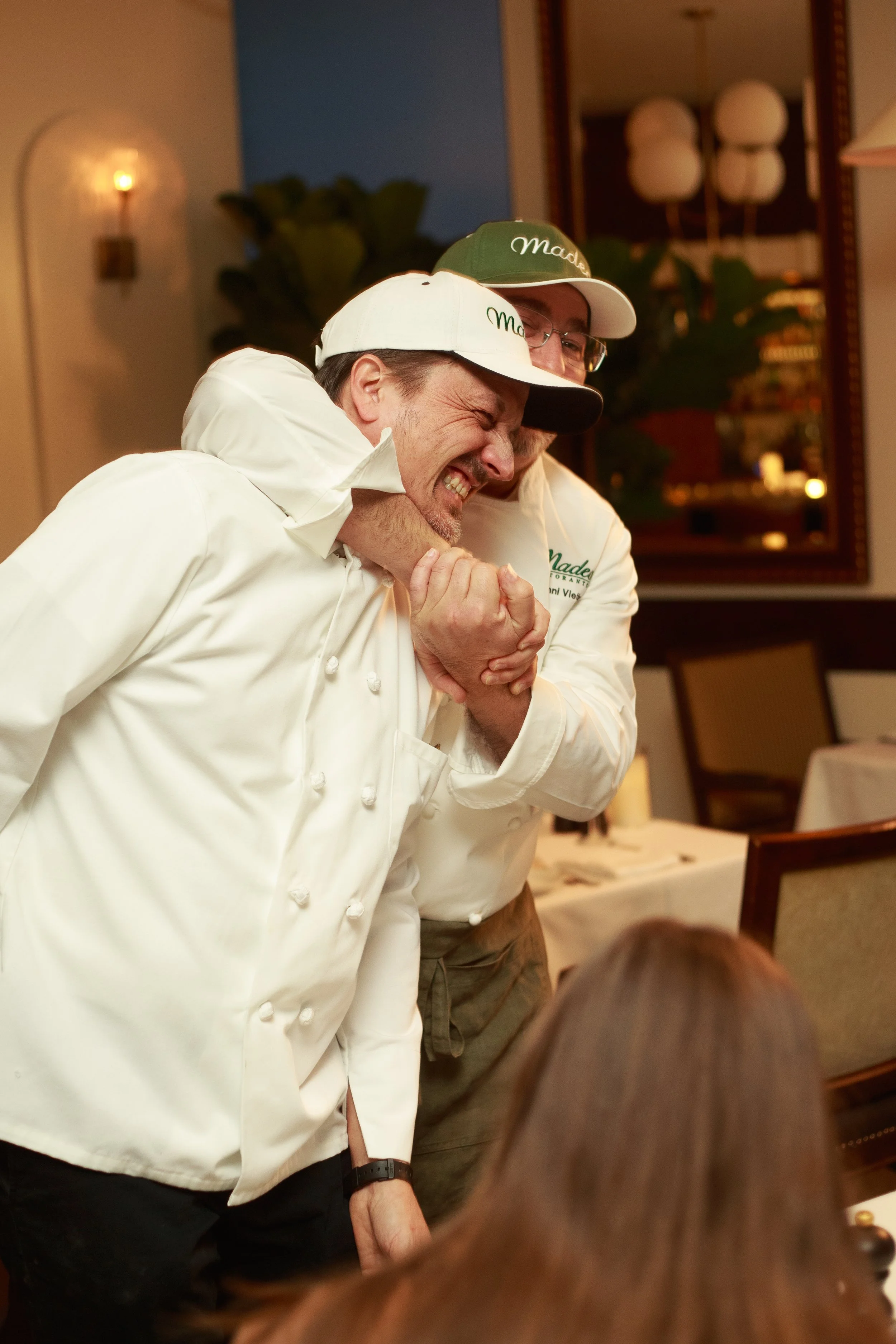 A chef is holding another man in a chokehold during a playful fight at a restaurant, with the chef grimacing and the other man appearing to struggle.