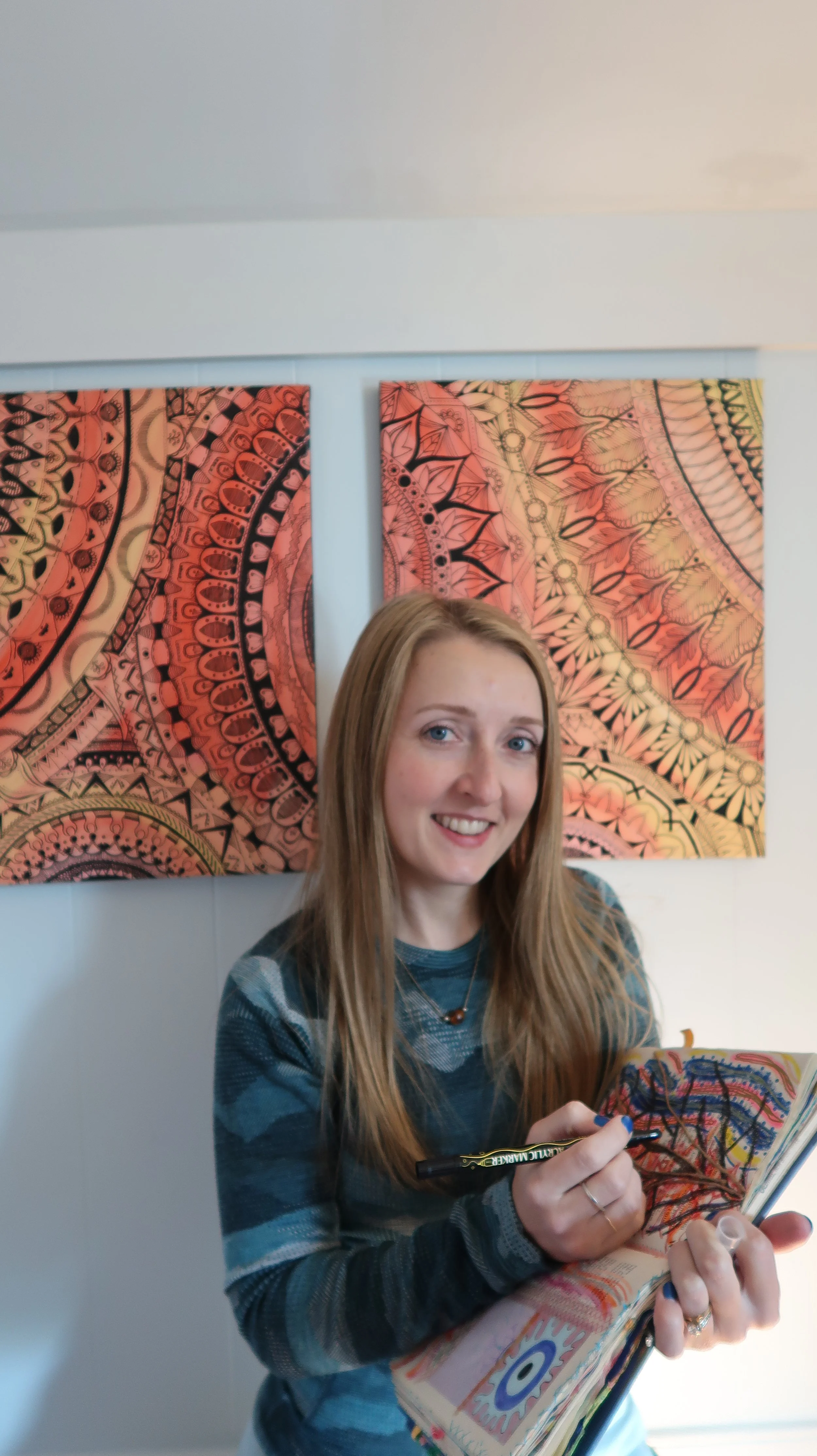 A woman with long blonde hair smiling and holding a book with colorful drawings, standing in front of two pink, mandala-like artworks on a white wall.