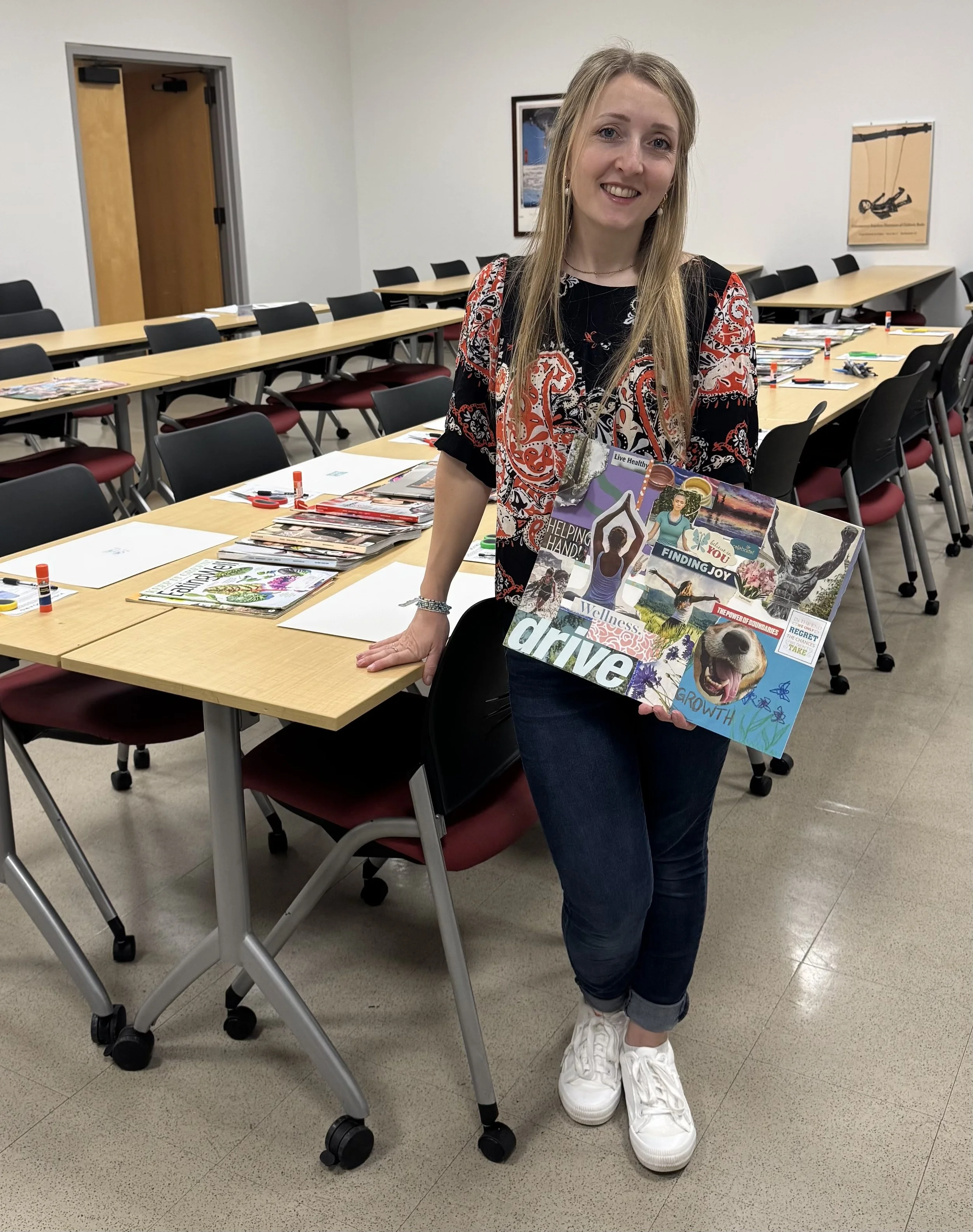A woman standing in a conference room holding a collage or poster with various images and words related to wellness, growth, and joy. The room has a long table with chairs, some magazines, and markers.