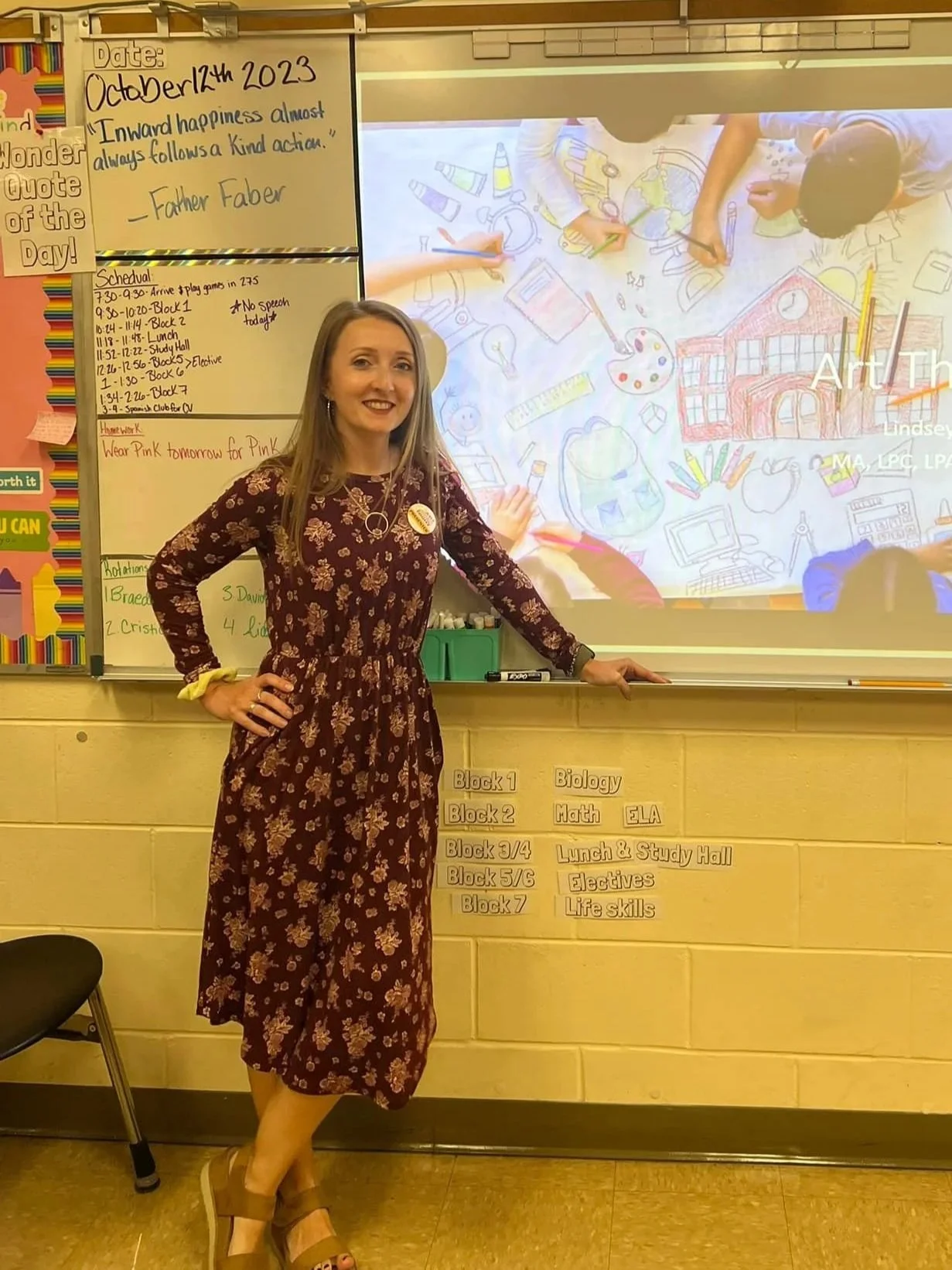 A teacher standing in a classroom in front of a whiteboard and a projector screen, smiling and posing for the photo. The whiteboard has a date and a quote, and the projector screen displays children drawing on a large piece of paper.