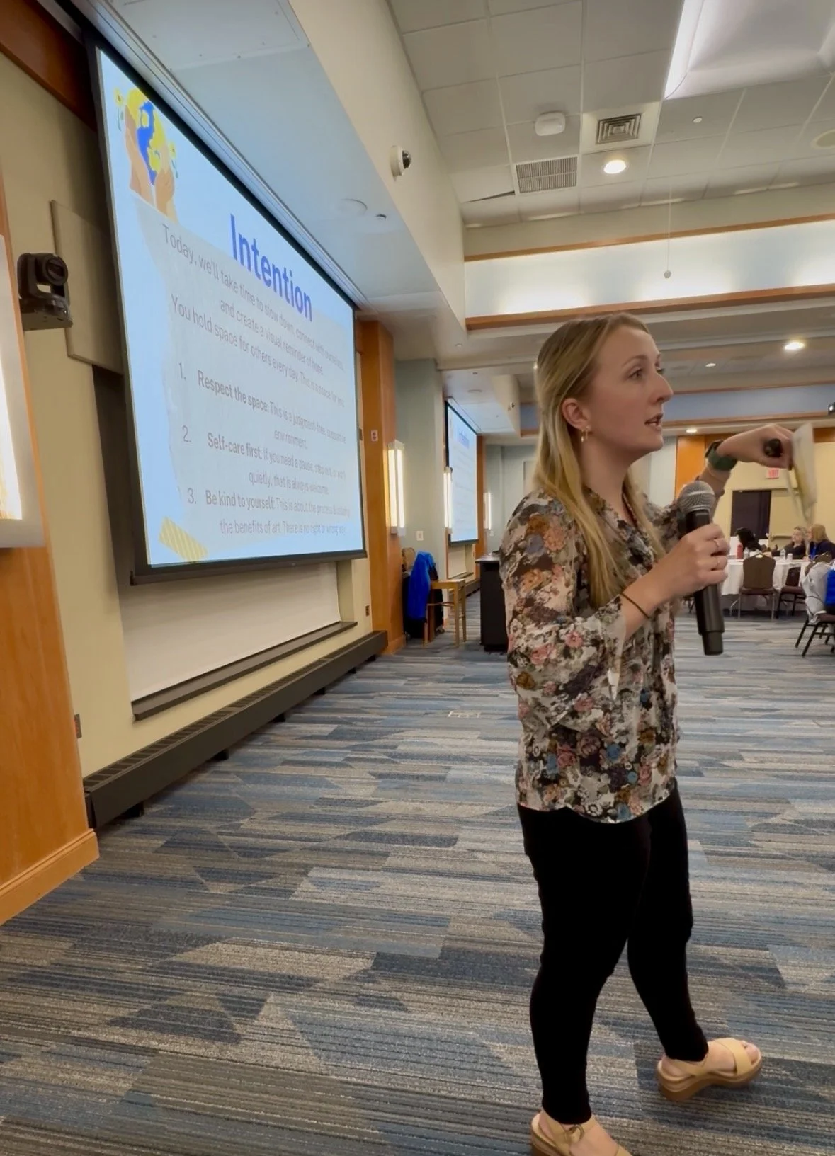 A woman with long blonde hair holding a microphone and standing in front of a large screen displaying a presentation on attentiveness. She is wearing a floral blouse, black pants, and beige shoes, in a conference room with tables and chairs, and several attendees in the background.