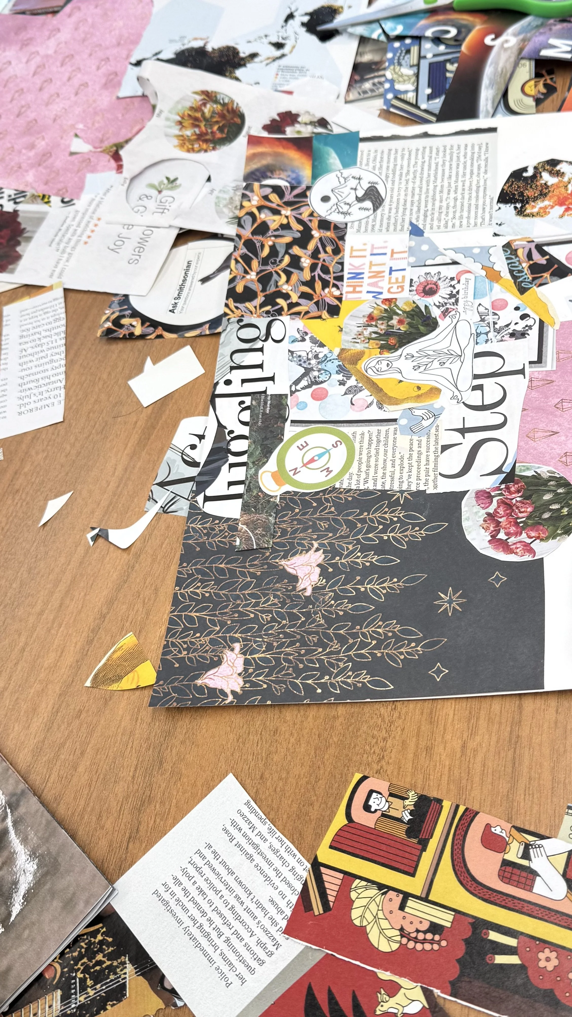 A cluttered table with scattered magazines, paper cutouts, stickers, and illustrations, including floral and abstract designs, with some pink fabric or paper partially visible on the edge.