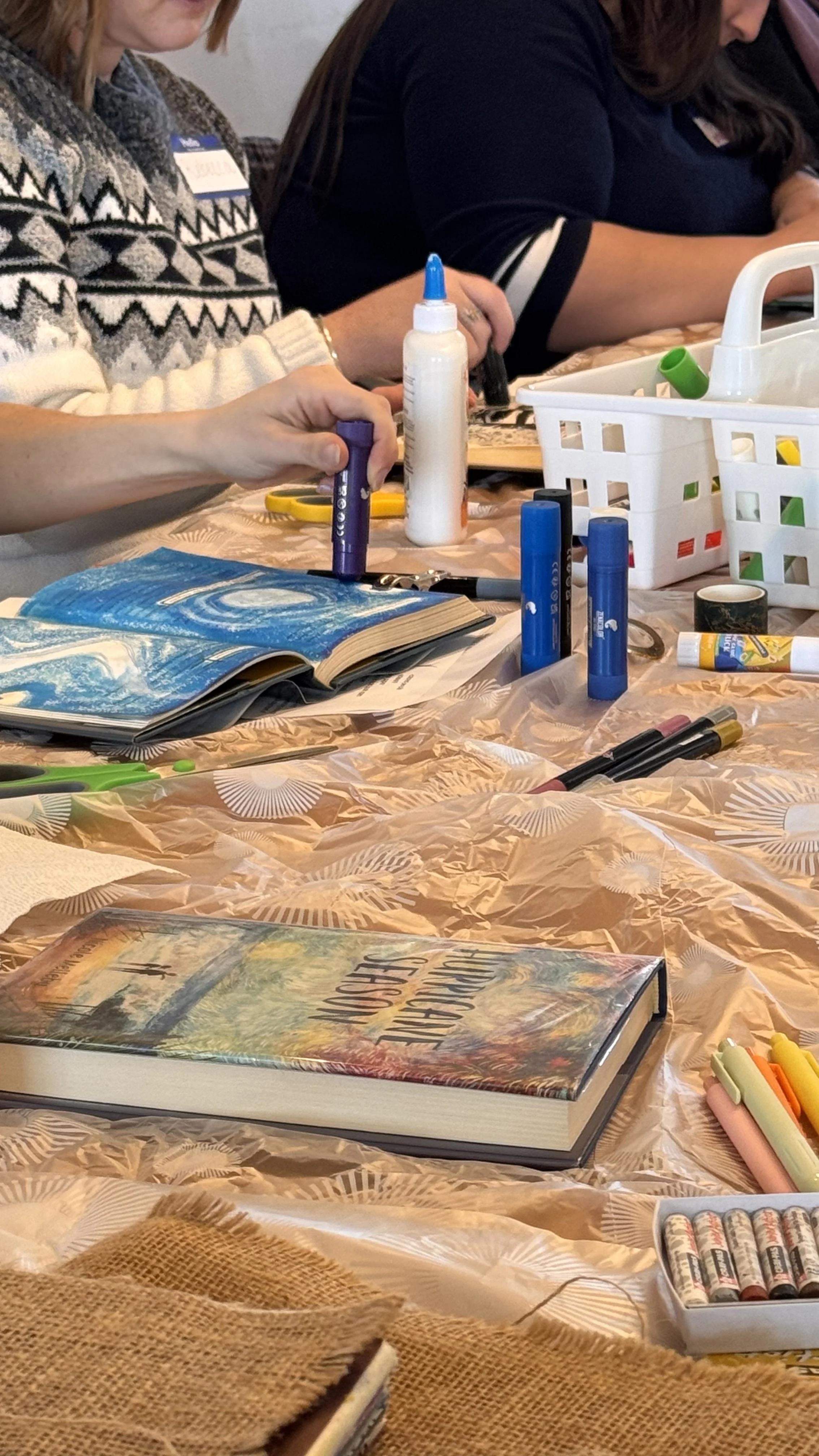 People painting on a table with paint bottles, brushes, and a paper plate, with a child's drawing in the foreground.