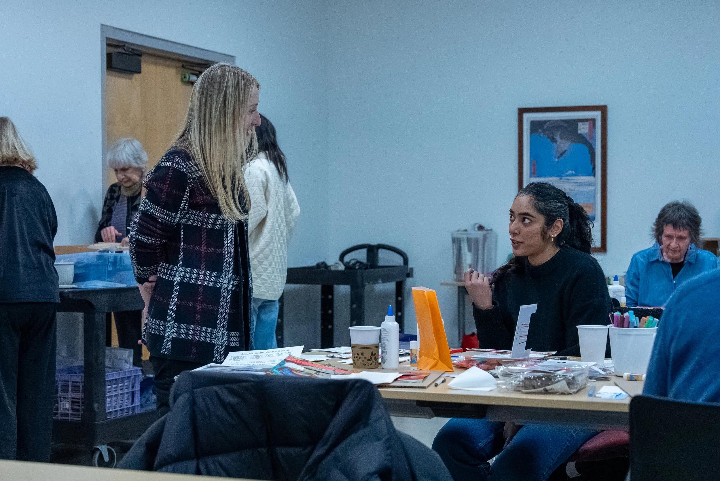 Two women are having a conversation at a table in a room with other people. The seated woman has dark hair tied back, and the standing woman has long blonde hair. The table has various papers, markers, and a small orange bag.