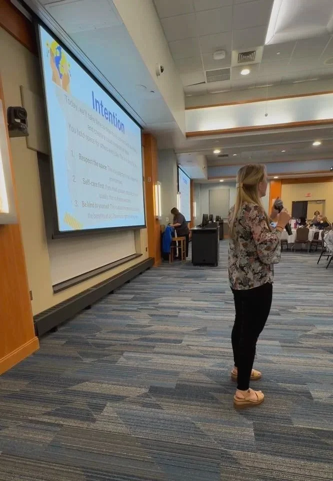 A woman giving a presentation in a conference room, standing near a large projection screen displaying a slide titled 'Intentions' with three bullet points.