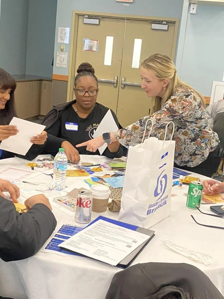 A group of women gathered around a table engaged in discussion, with papers, cans, a water bottle, and a large white branded bag on the table.