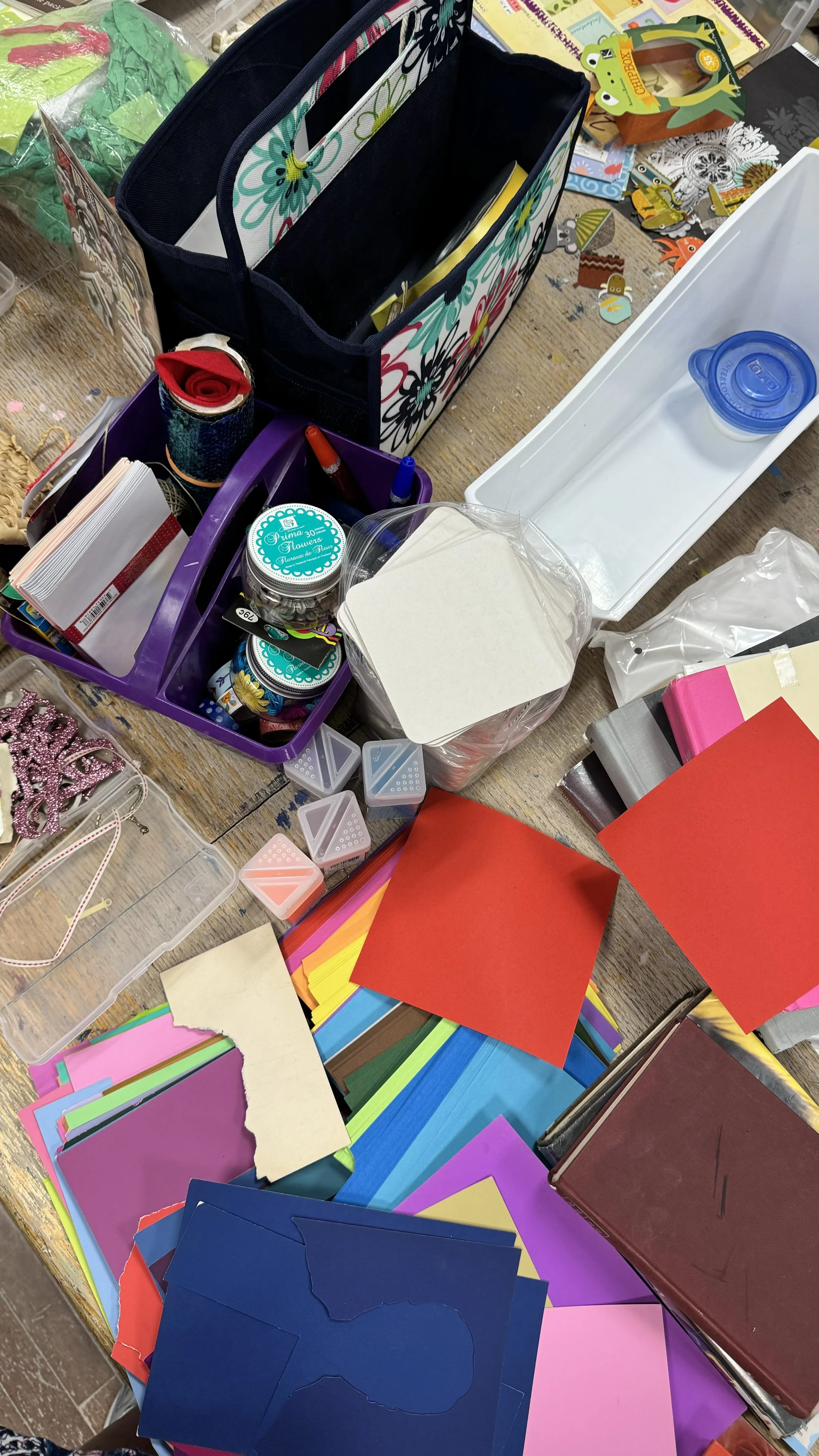 Collection of craft supplies on a table, including colored paper, stickers, decorative tape, and small containers for organizing craft materials.