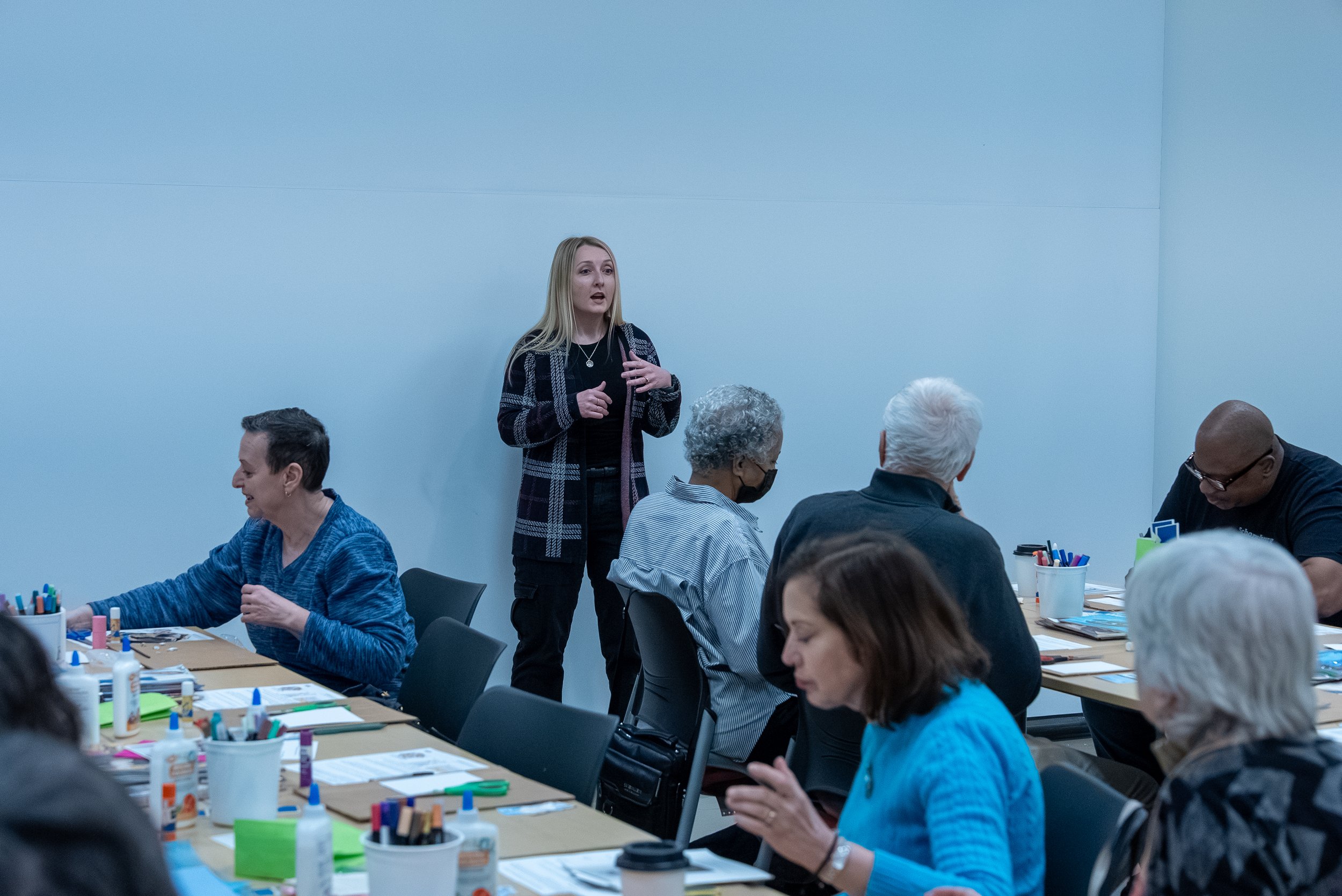 An art studio filled with various paintings, including a large mural of twisted trees on the left wall. Paint supplies, bottles, brushes, and canvases are arranged on tables in the center. Several completed artworks and paintings are leaning against the walls and on easels. Fluorescent lights illuminate the room.
