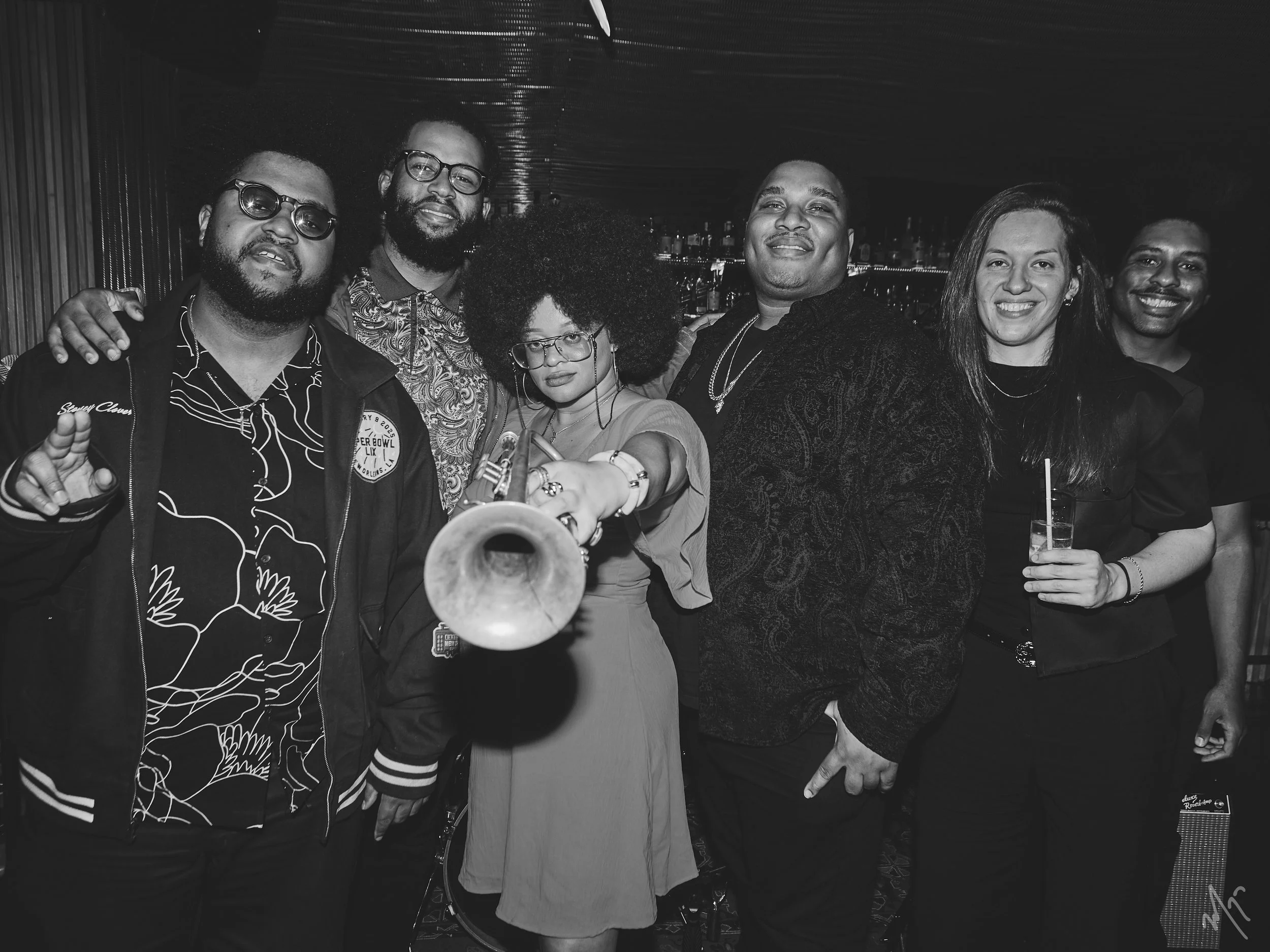 Group of seven diverse people posing together in a bar or nightclub, Tatiana Tate holding a trumpet, others smiling with drinks, all dressed casually.