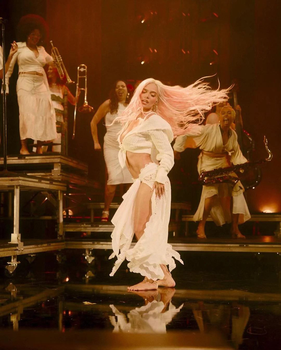 A woman with long pink hair dancing barefoot on stage, surrounded by musicians and backup singers in white clothing, with warm stage lighting and a reflective floor.