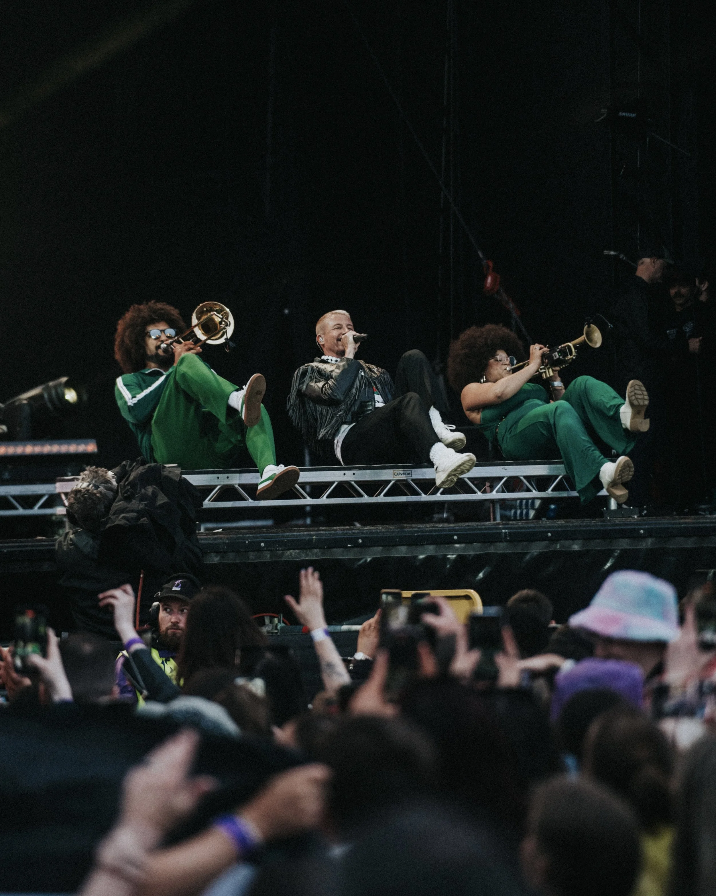 Band performing on stage with three musicians playing instruments, audience cheering and taking photos.