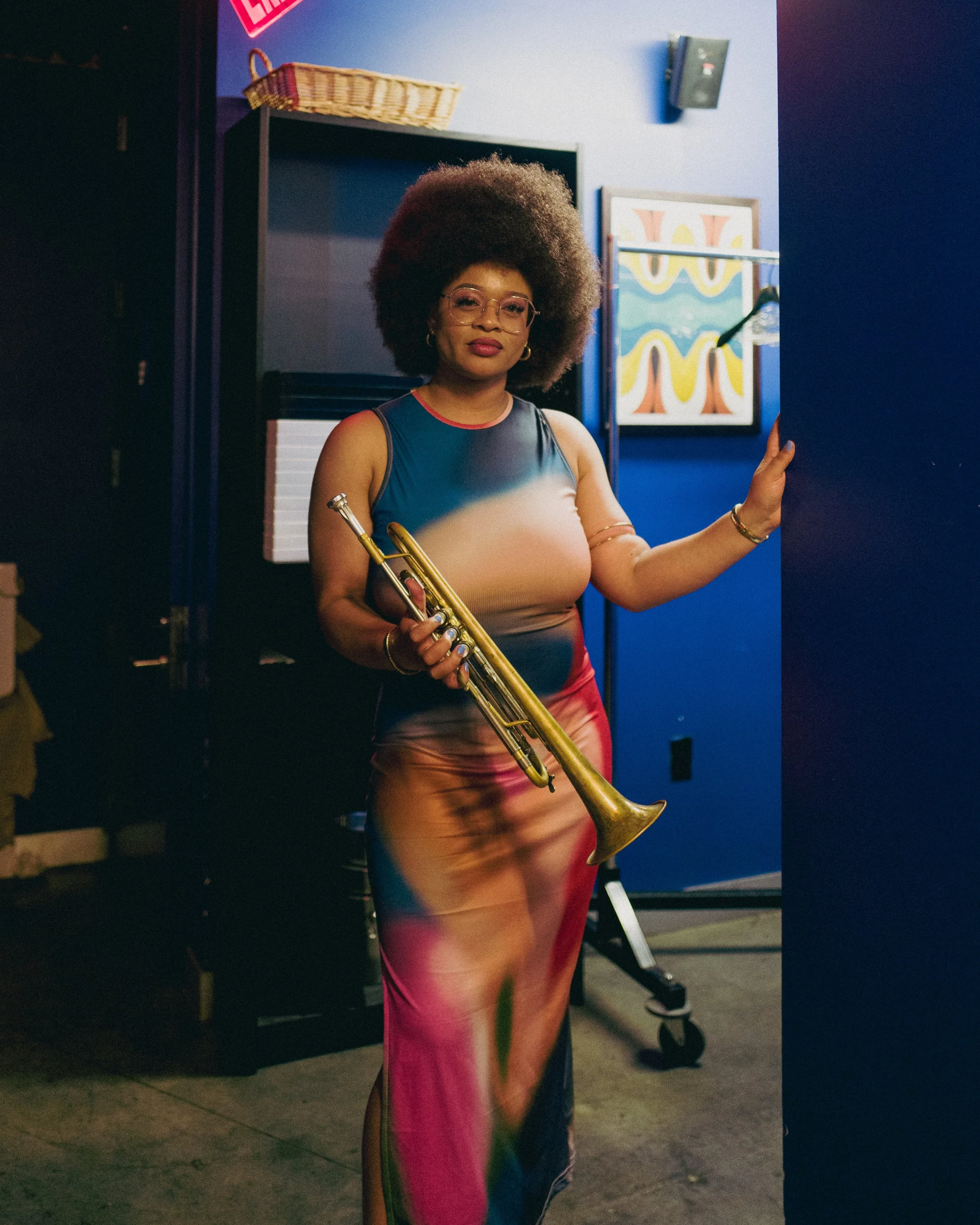 A woman with an afro hairstyle and glasses holding a small trumpet, standing in a colorful room with blue walls and artwork.