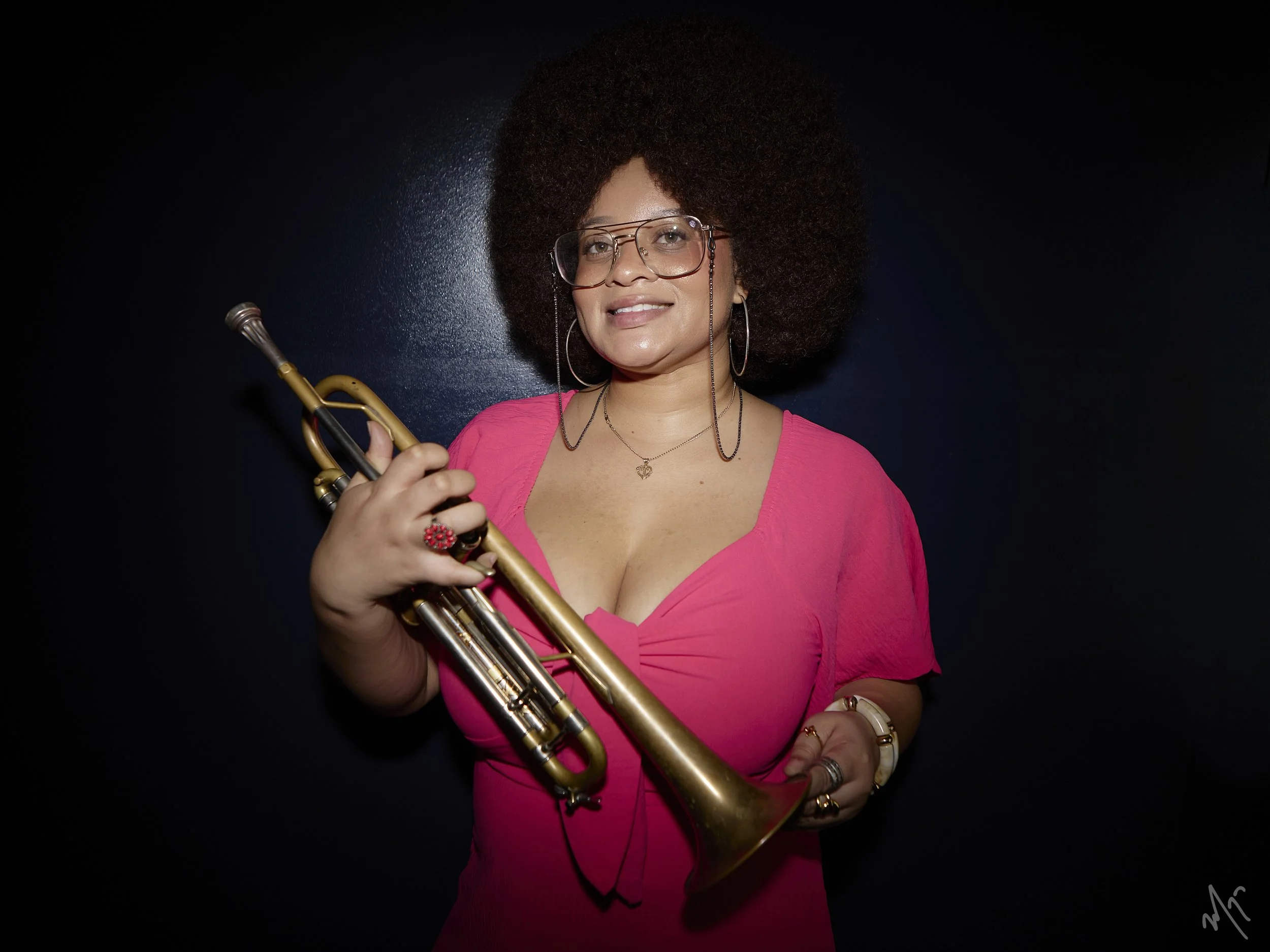 Tatiana Tate wearing glasses, a pink dress, and jewelry, holding a trumpet against a dark background.
