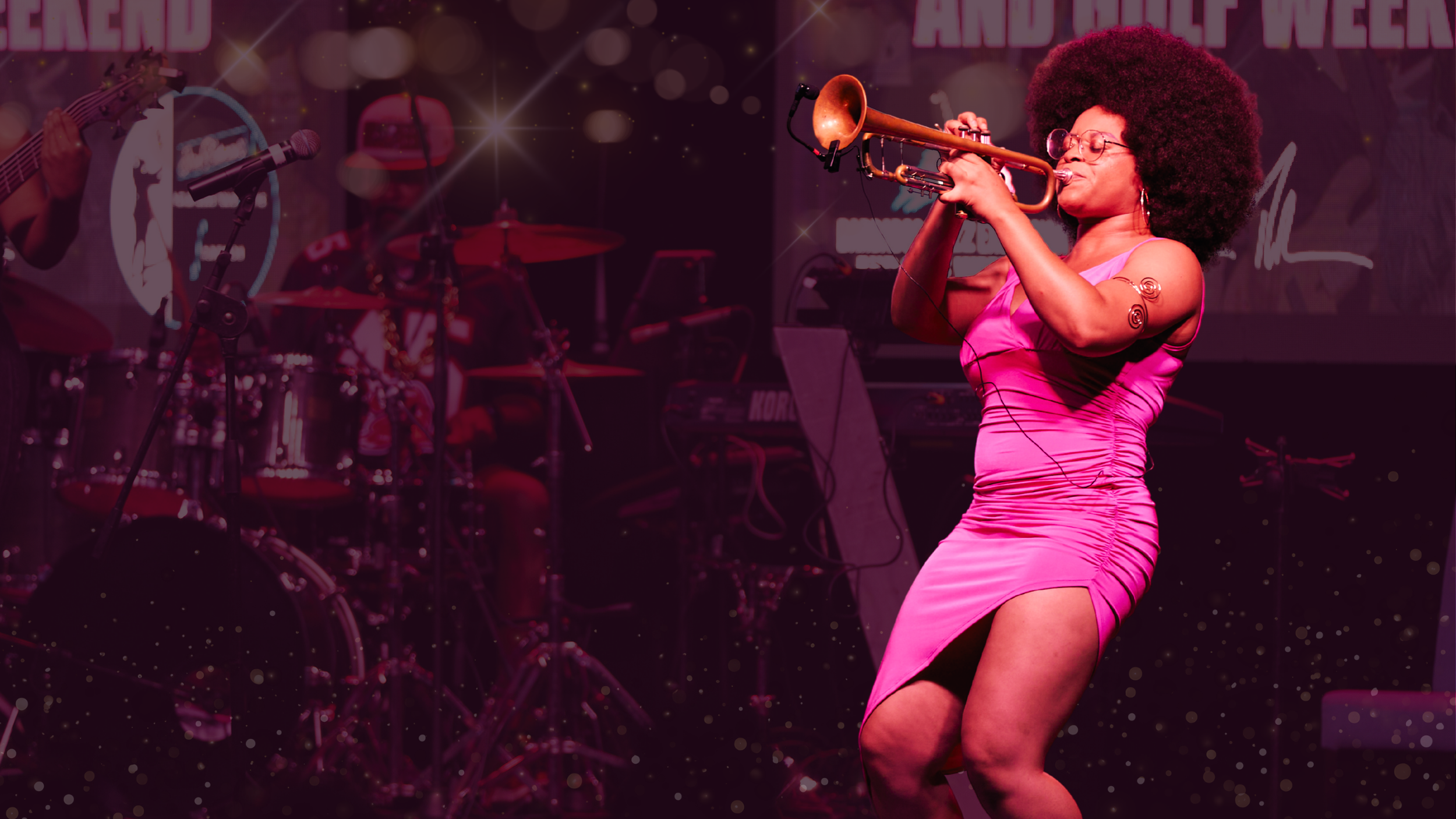 A woman in a pink dress playing a trumpet on stage with a band, with a heart-topped tattoo on her arm and an afro hairstyle.