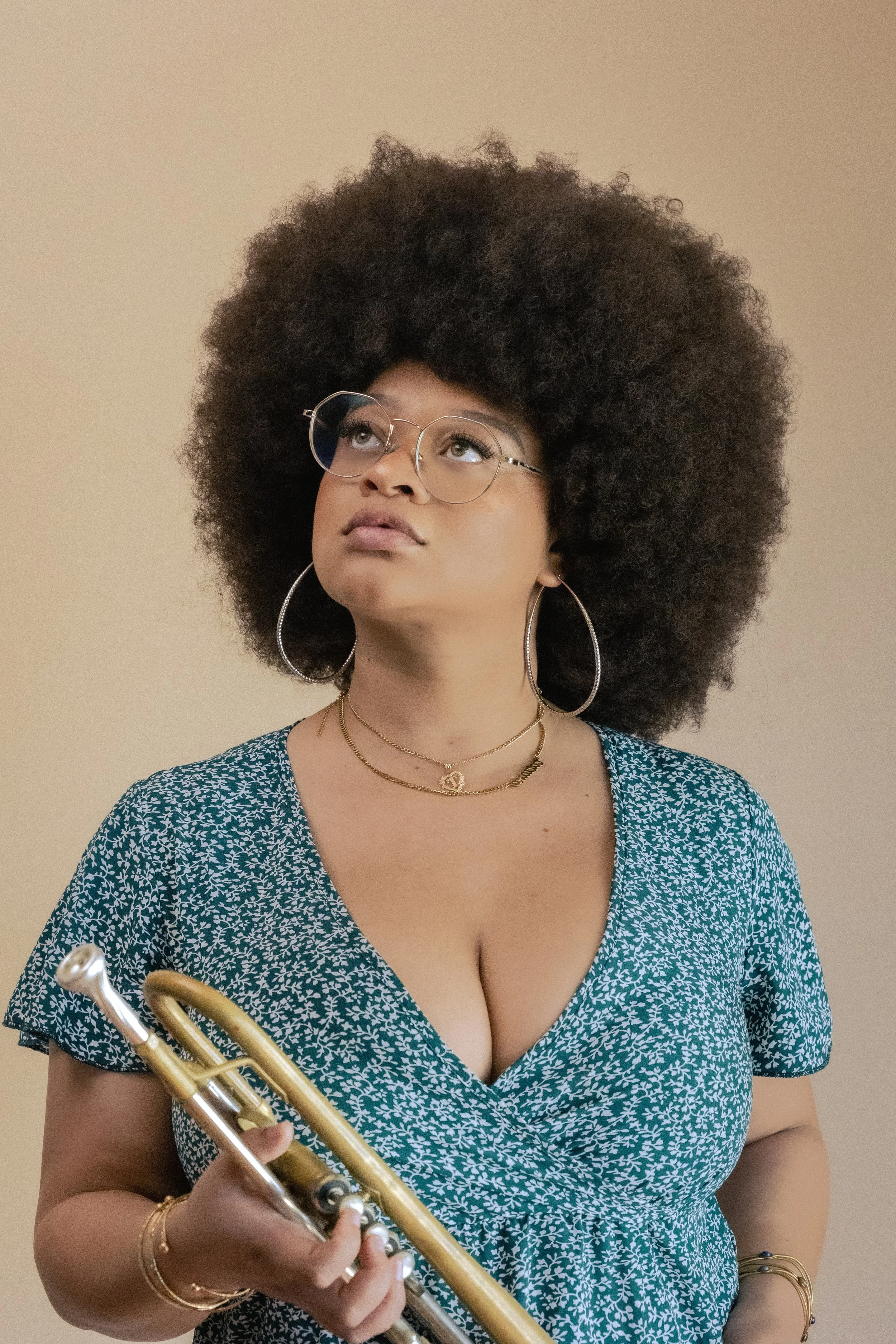 A woman with an Afro hairstyle, wearing glasses, hoop earrings, layered necklaces, and a blue patterned dress, holding a trumpet with a neutral background.