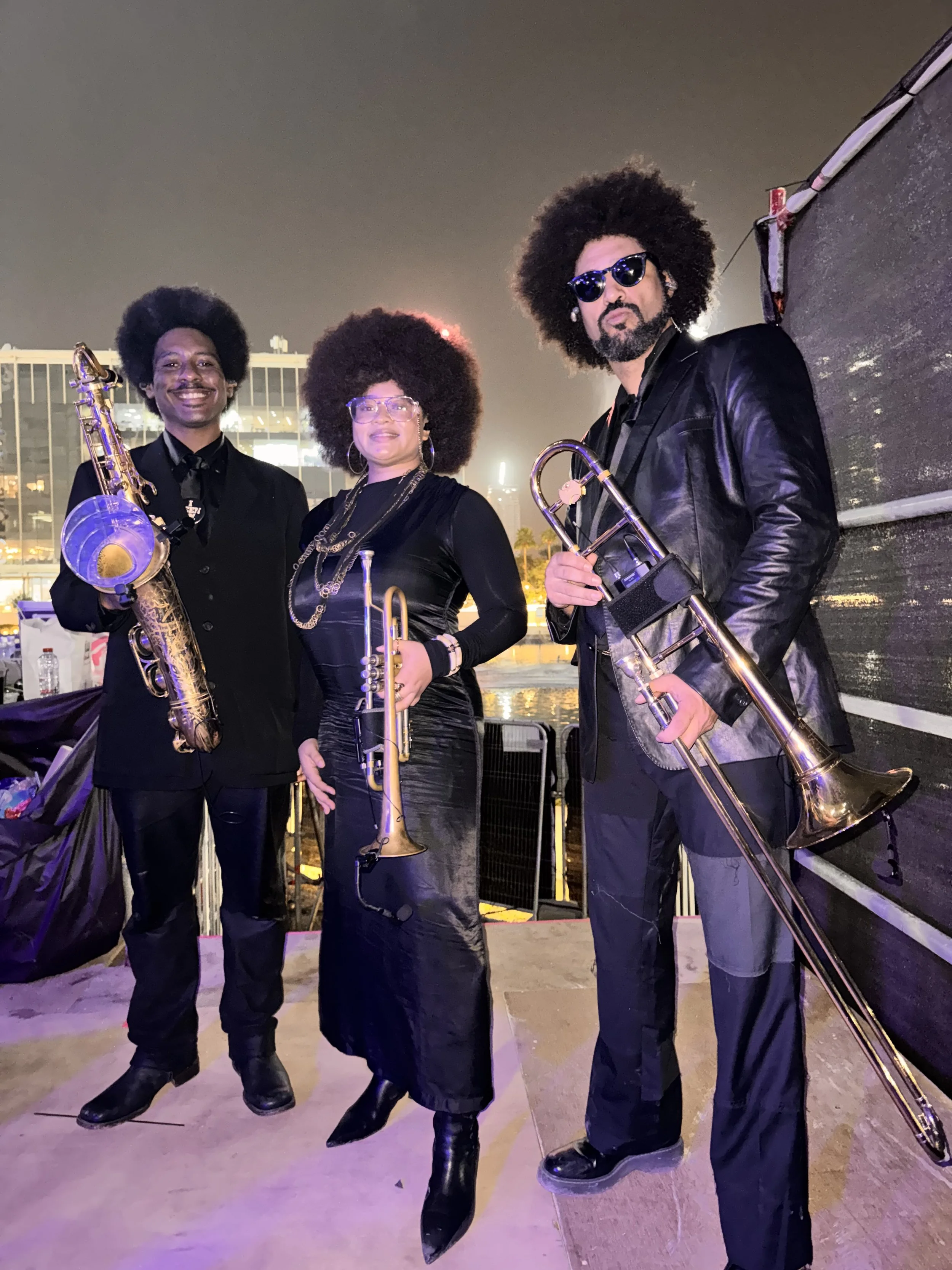 Three musicians with large afro hairstyles holding brass instruments, dressed in black, on an outdoor stage at night with city lights in the background.