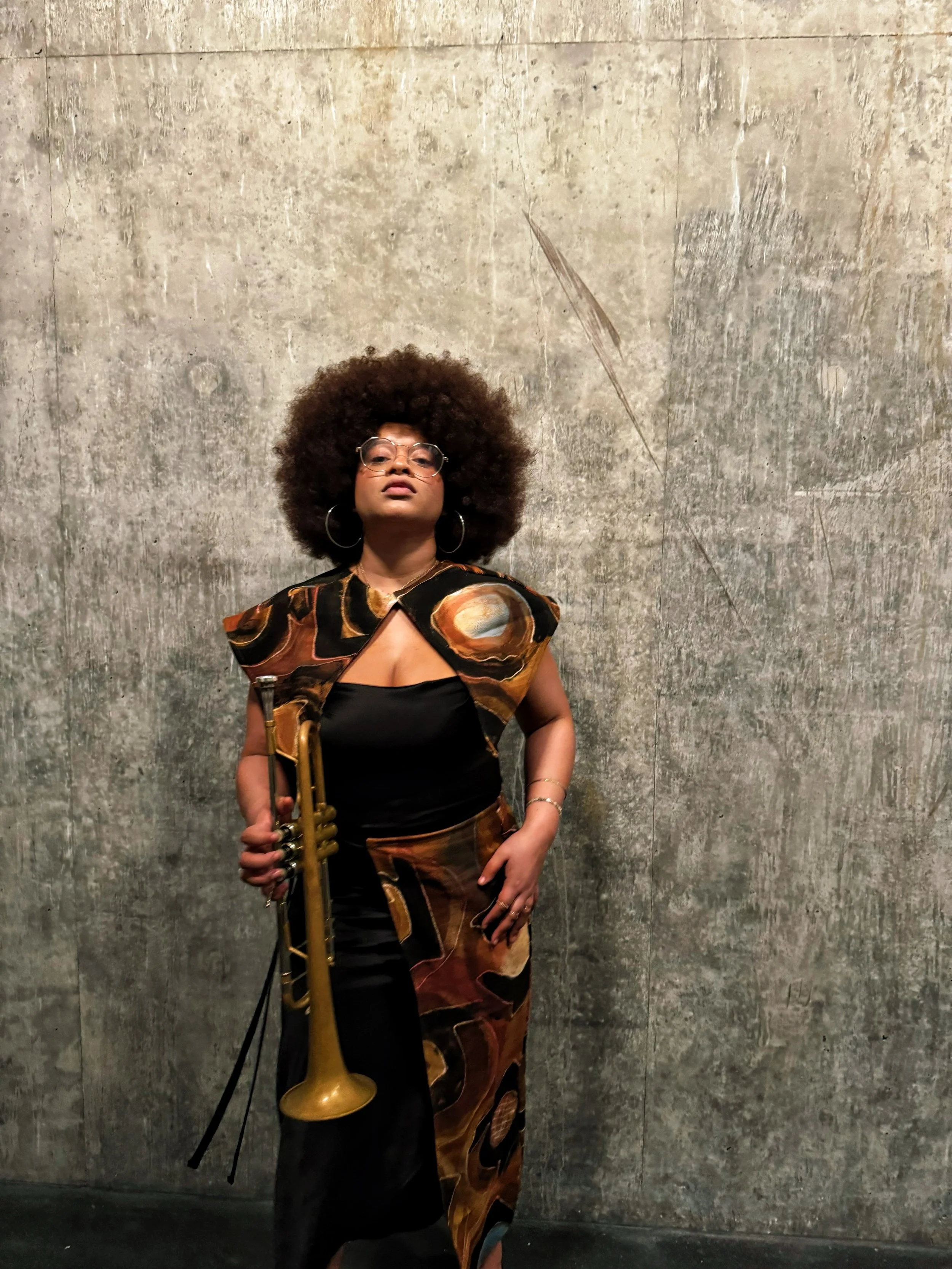 Tatiana Tate holding a trumpet, standing against a textured concrete wall.