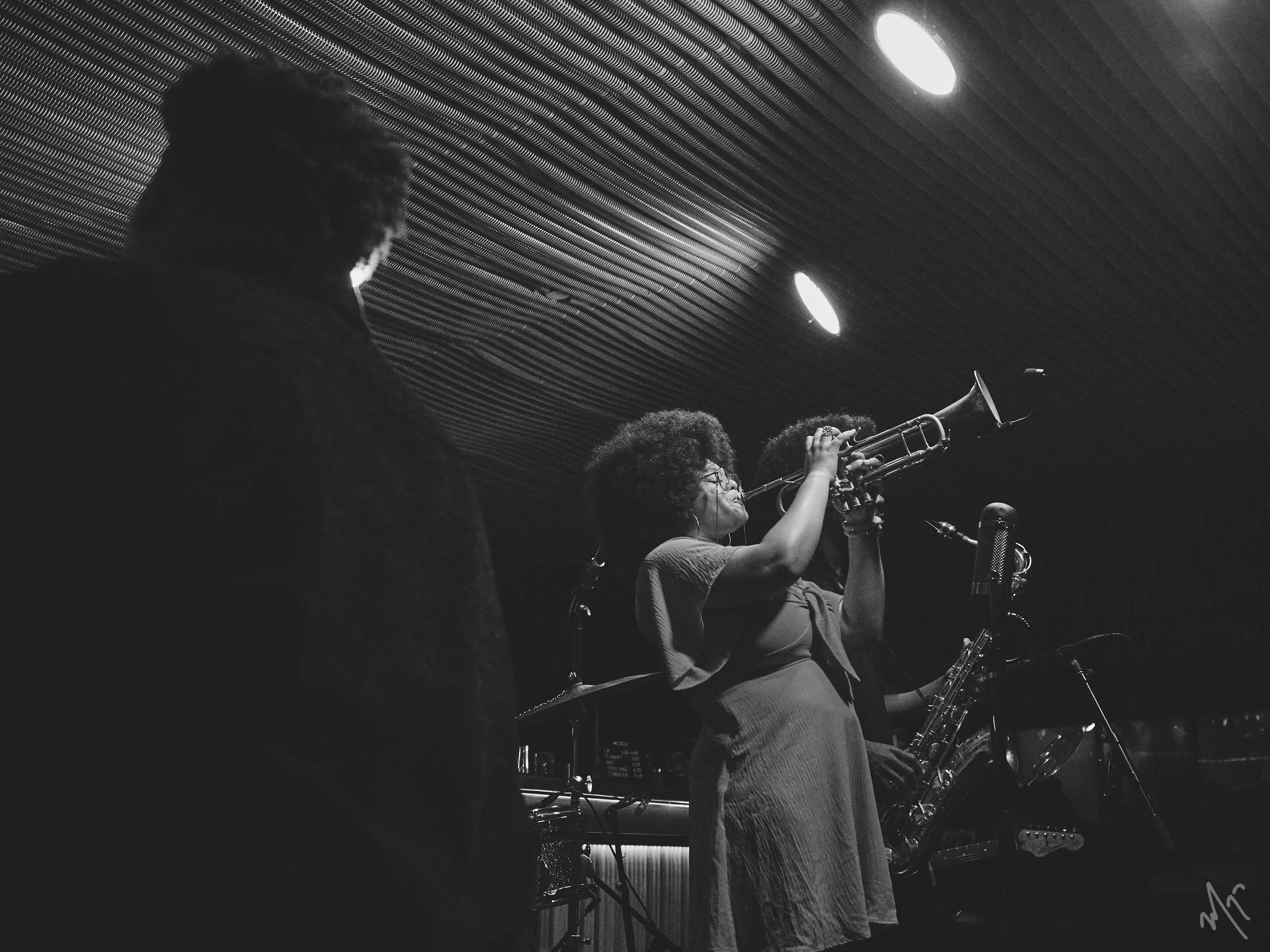 Black and white photo of Tatiana Tate playing the trumpet while standing next to a microphone, surrounded by other musicians. A person on the left is watching the performance in a dimly lit room with a textured ceiling.