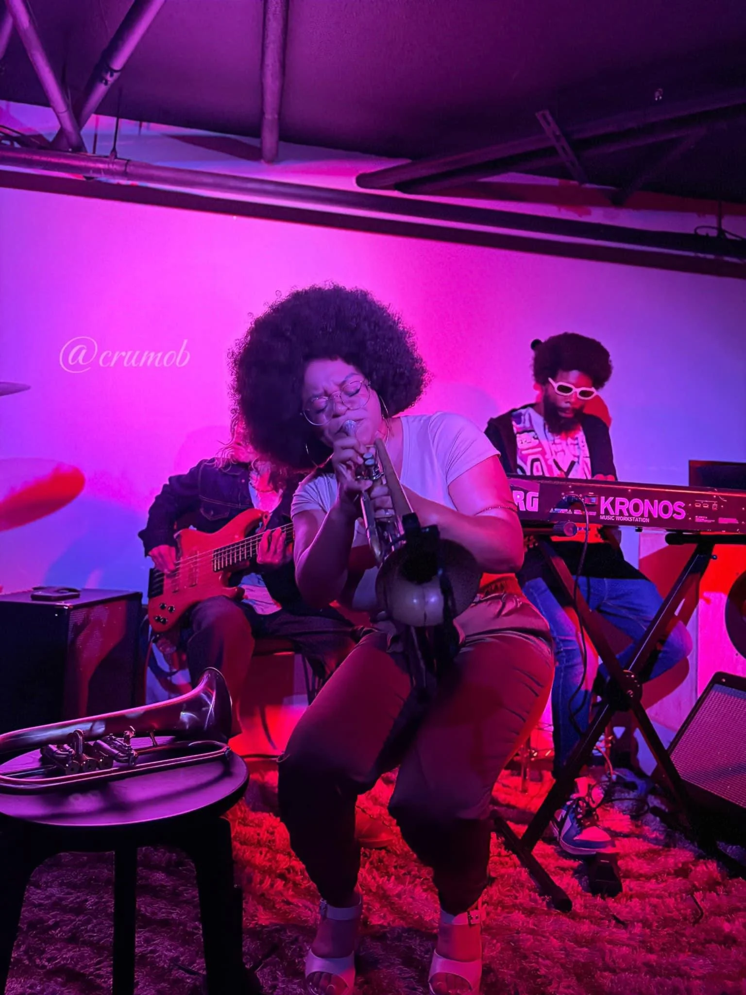 A woman passionately singing into a microphone while playing a saxophone, with two male musicians in the background playing guitar and keyboard. The scene is illuminated with pink and purple lighting, and a small table with a trumpet and a tuner is in the foreground.