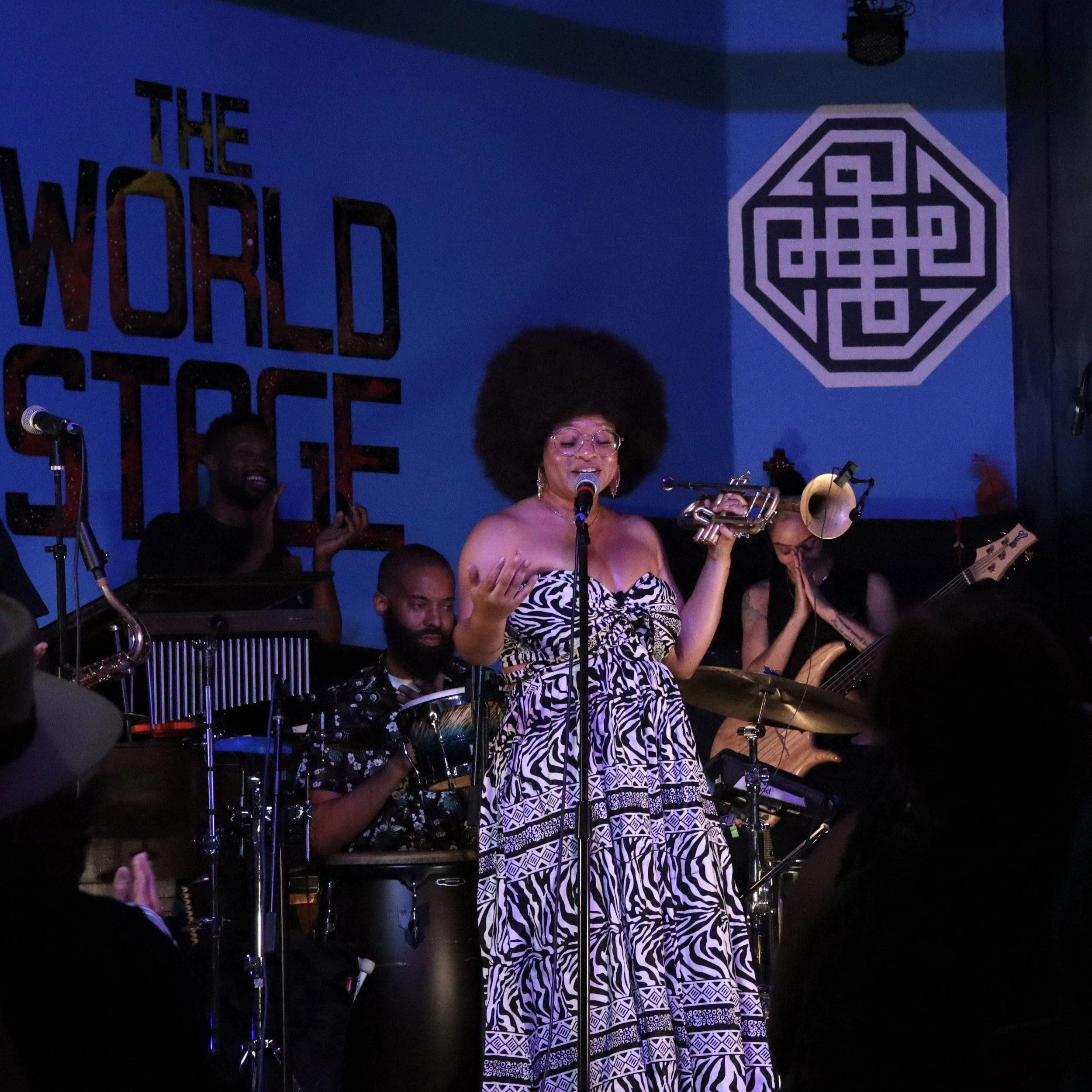 Tatiana Tate singing into a microphone on stage while holding a trumpet, with a band playing behind her in a venue with a blue background and the words "The World Stage" on the wall.