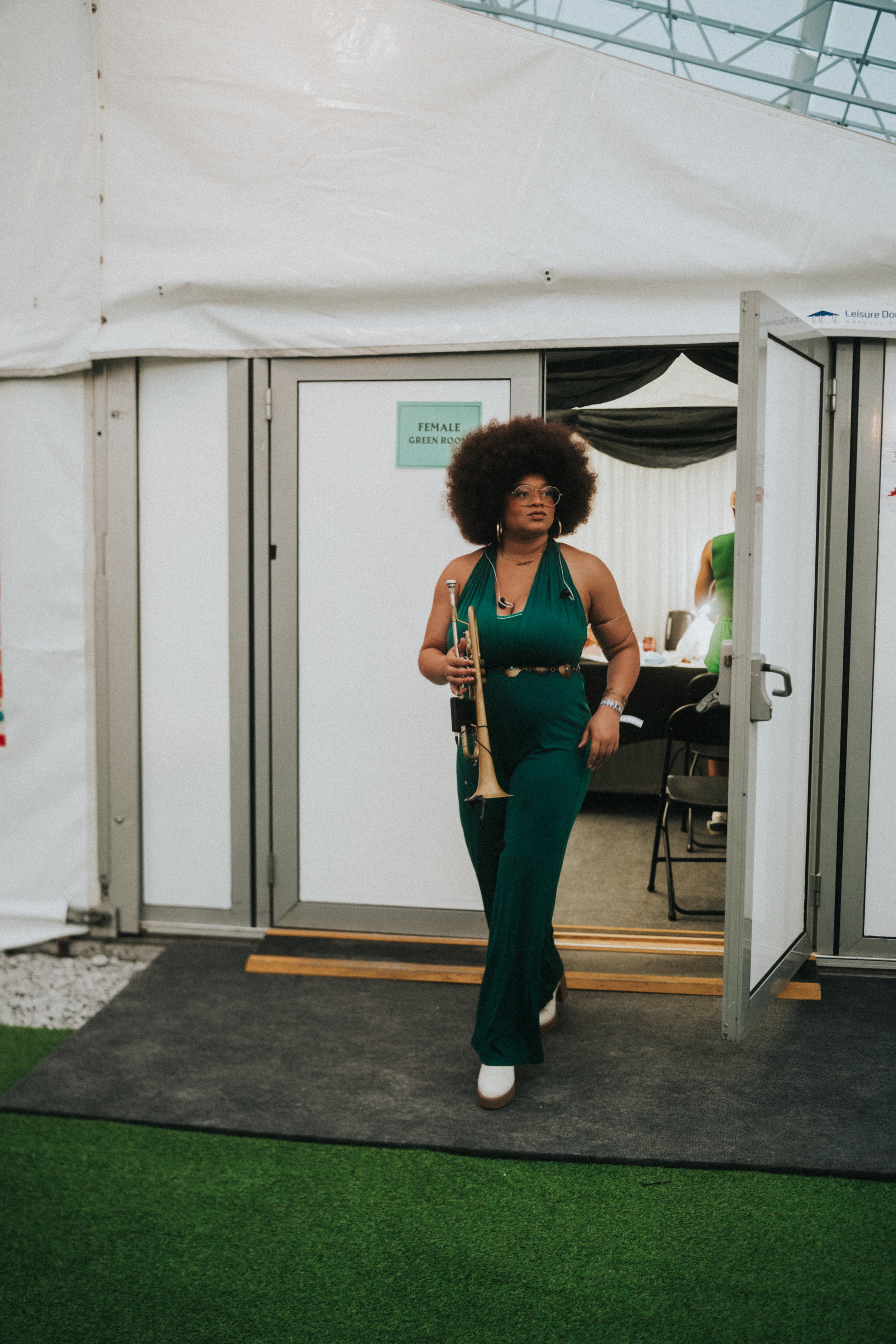 Tatiana Tate walking out of a tent, holding a trumpet, dressed in a green jumpsuit, with a black belt, white shoes, and accessories, emerging from a door labeled 'Female Green Room'.