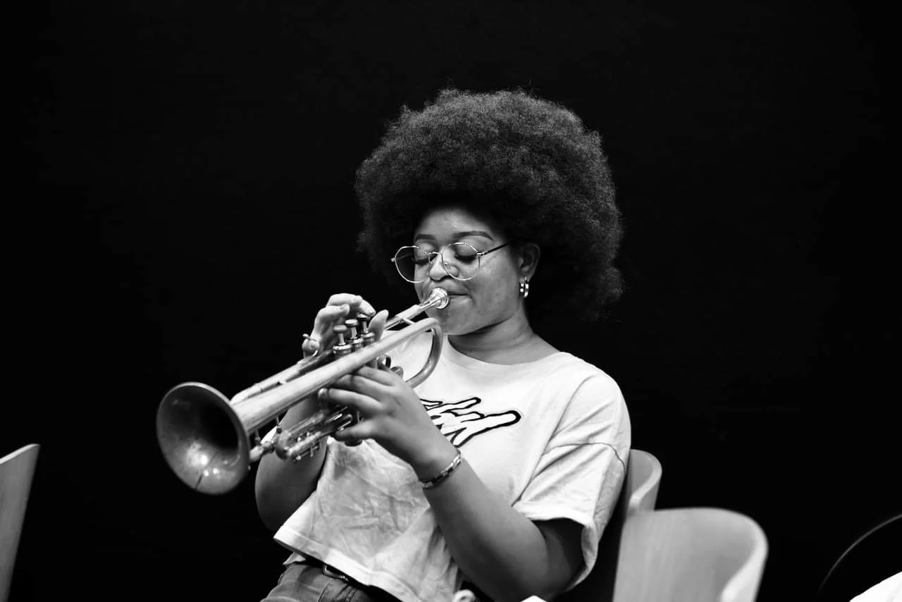 Tatiana Tate playing a trumpet, wearing glasses, earrings, and a t-shirt, sitting against a dark background.