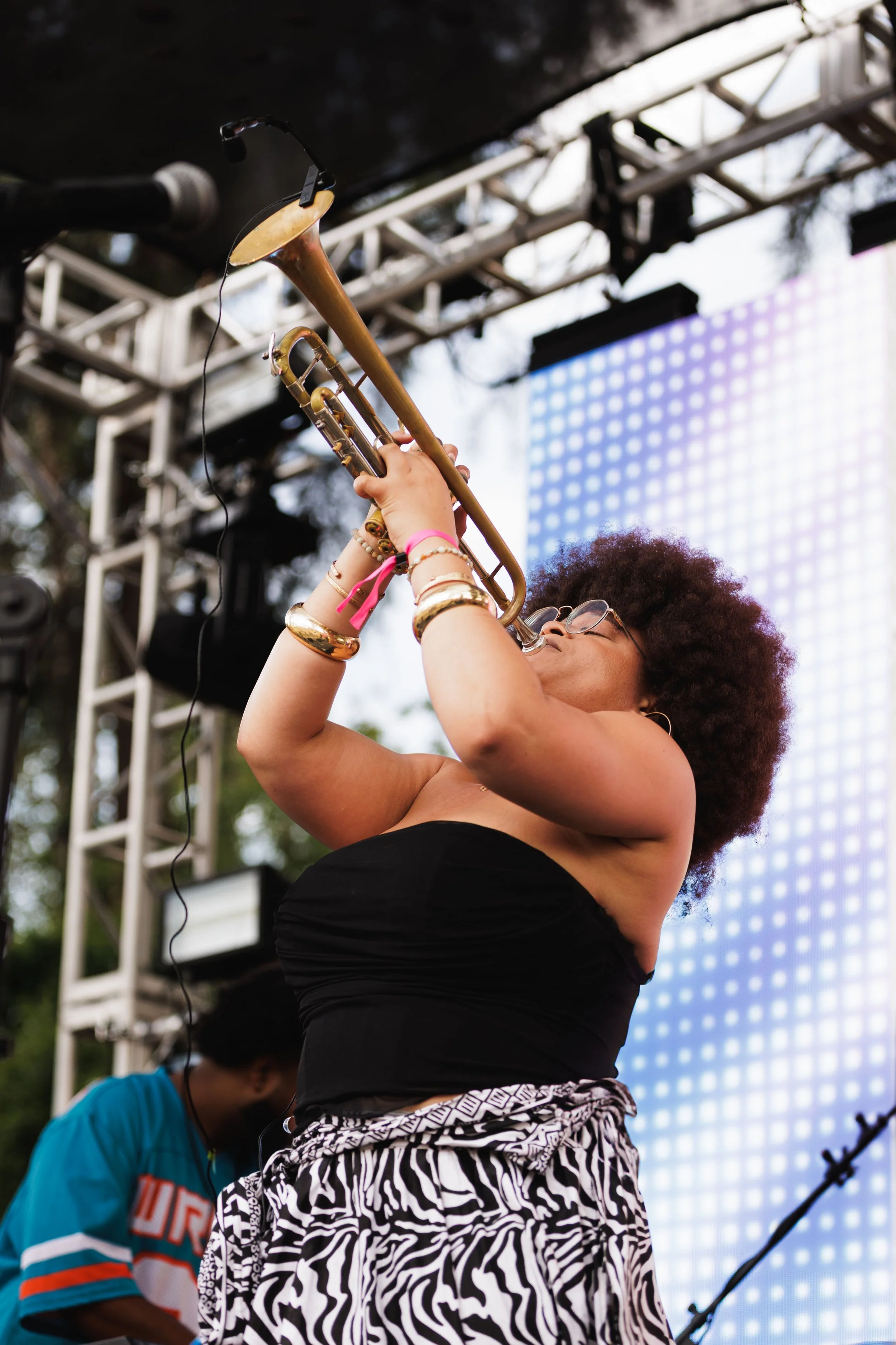 A woman with curly hair and glasses playing a trumpet on an outdoor stage, with a man in a sports jersey in the background.