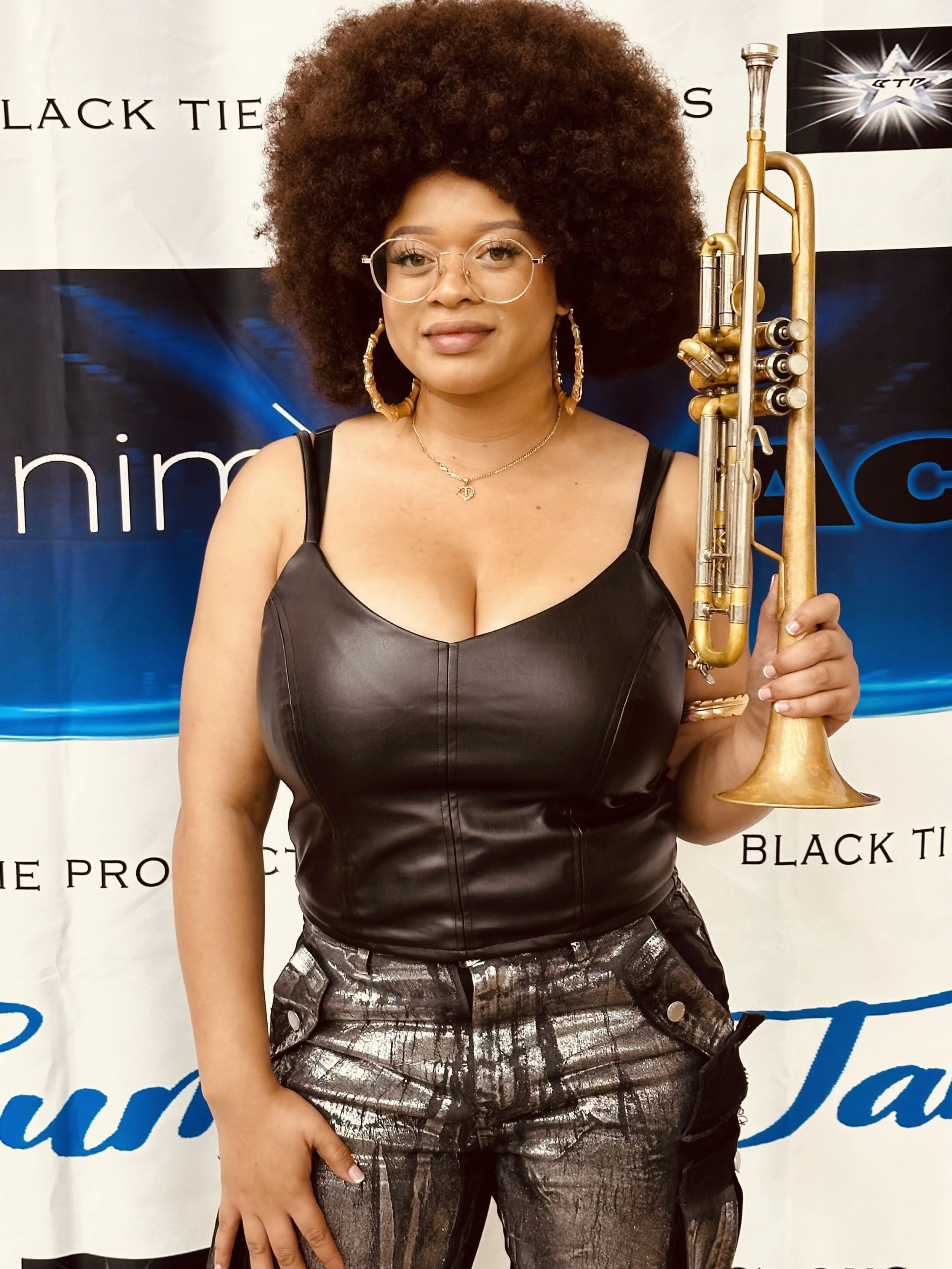 Woman with afro hairstyle, wearing glasses, hoop earrings, a black leather top, patterned pants, and holding a trumpet, standing in front of a backdrop with text.