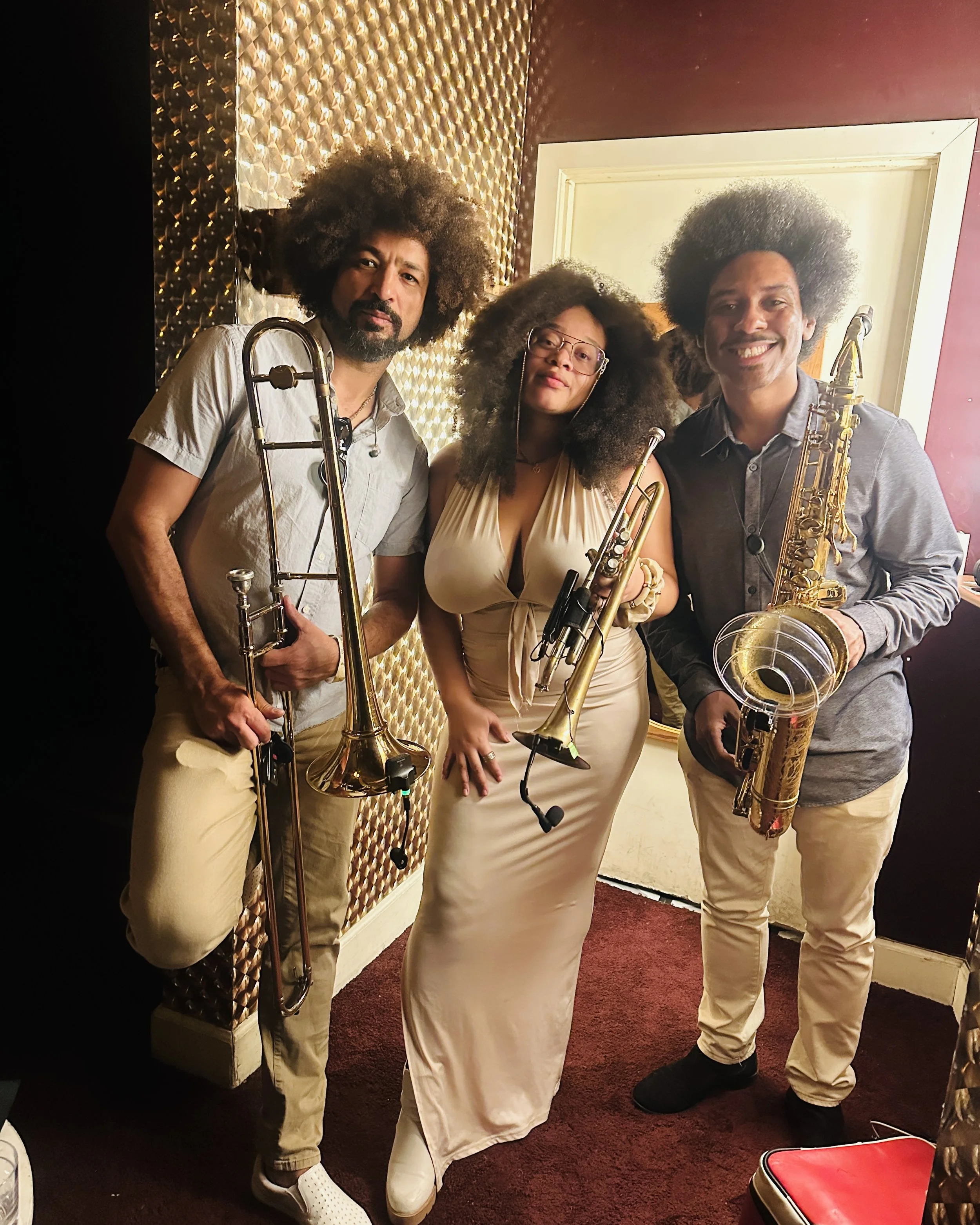 Three musicians with afro hairstyles holding brass instruments, standing in a room with gold and pink accent walls, posing for a photo.
