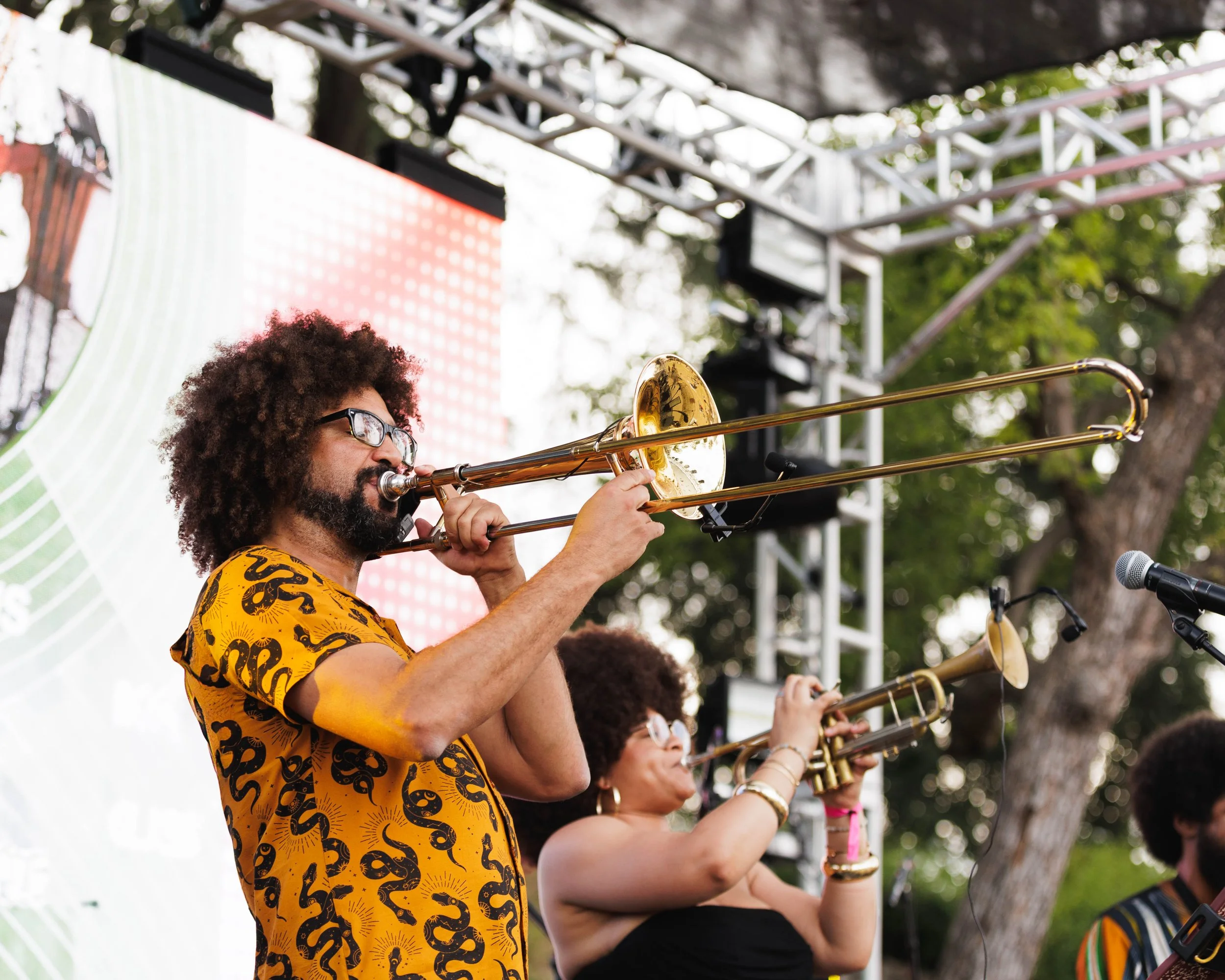 Musicians performing on stage outdoors, playing trombones, with a man wearing glasses and a yellow patterned shirt, and a woman with braids and jewelry, during a live concert.