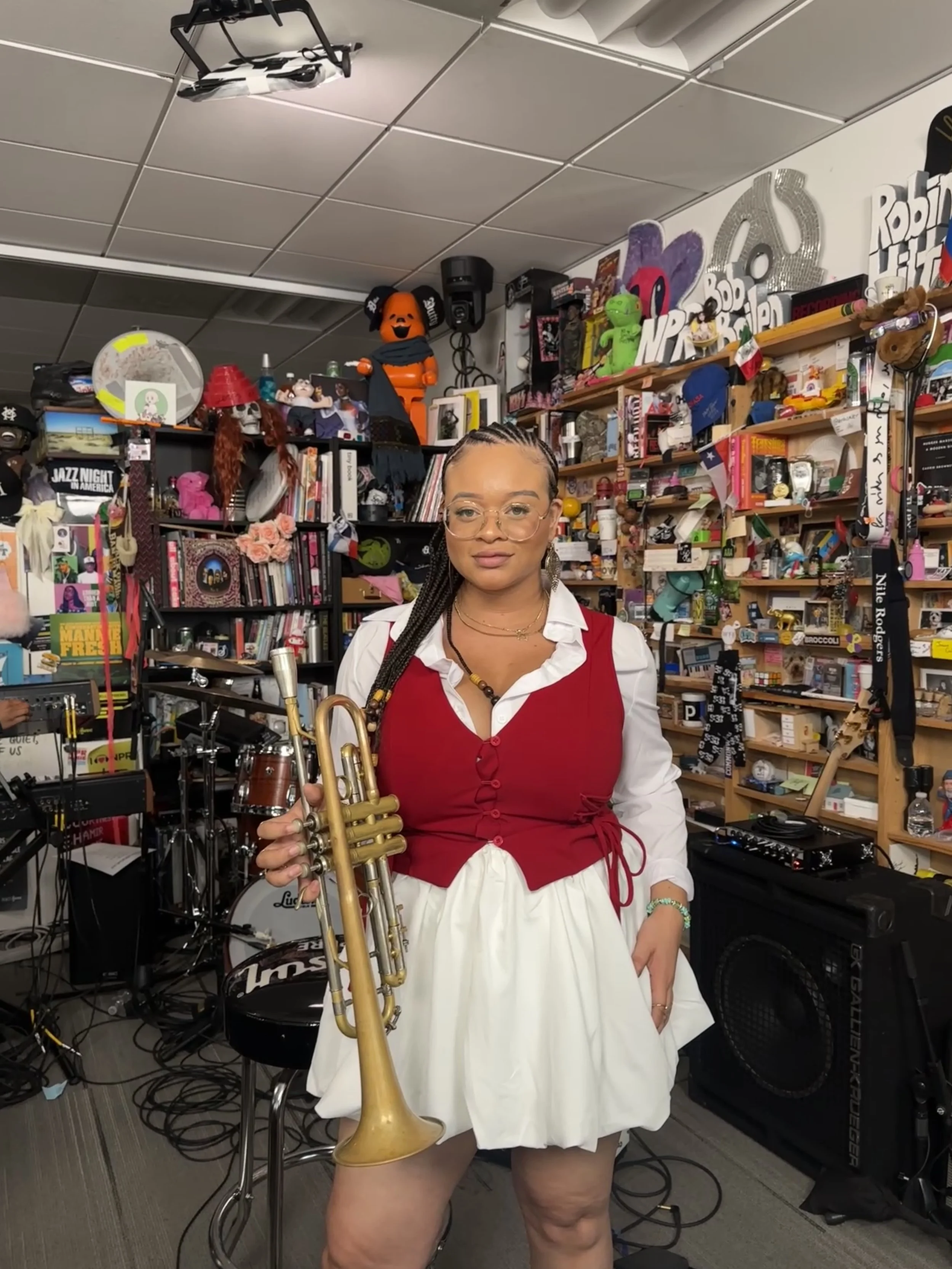 Tatiana Tate  holding a trumpet in a room filled with various toys, books, and decorations, with shelves full of colorful items and musical equipment behind her.