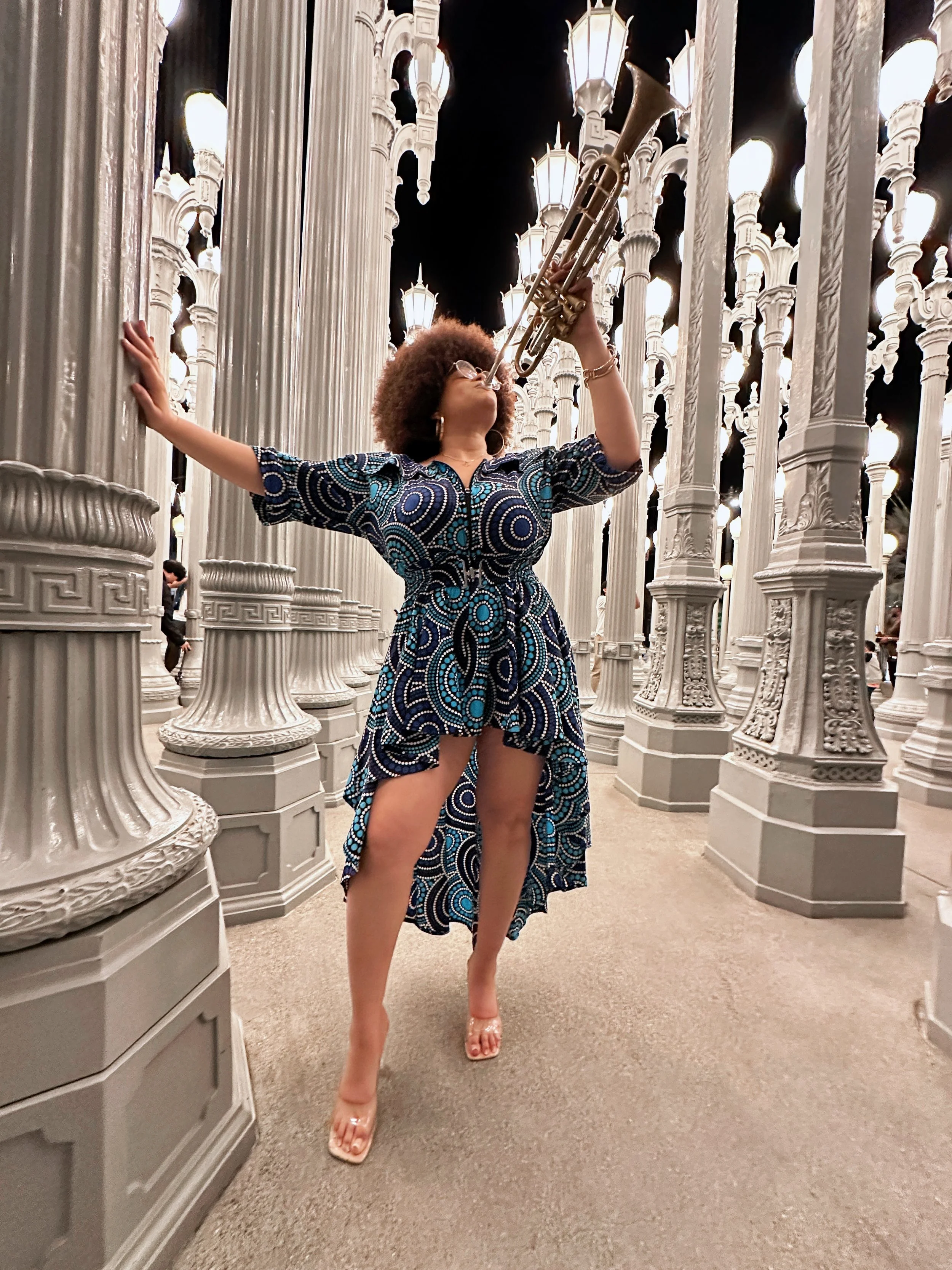 Tatiana Tate plays a trumpet in front of ornate white columns illuminated by large lights at night.