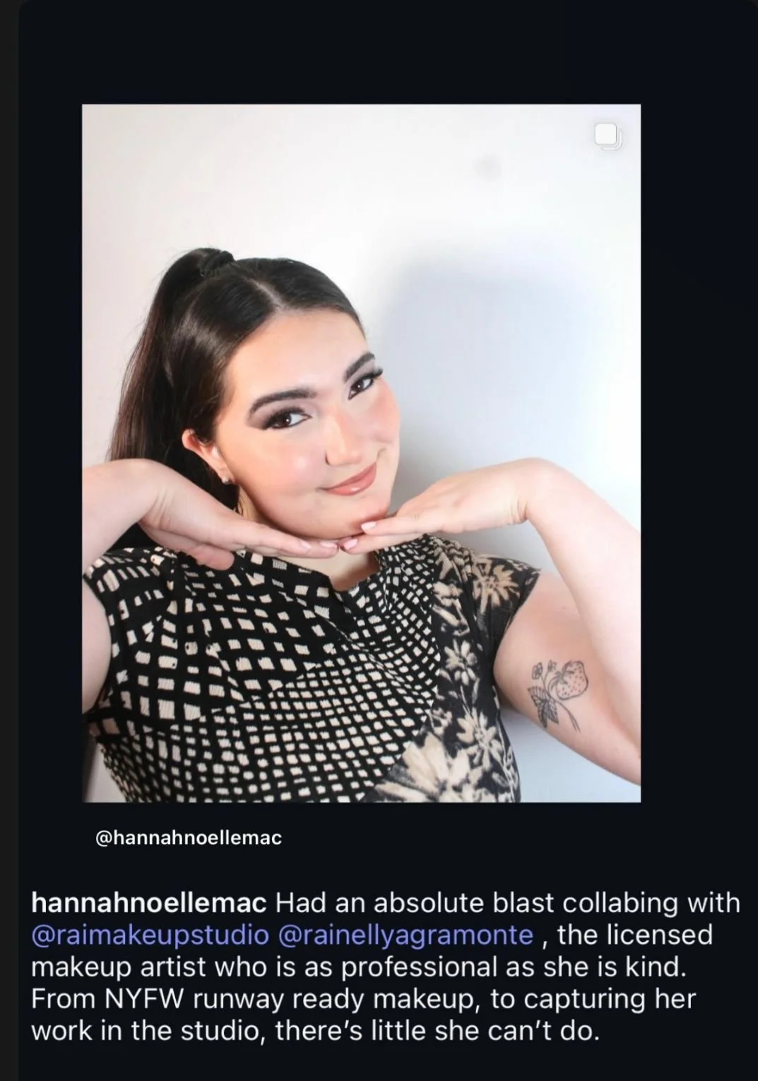 A woman with dark hair styled in a high ponytail, wearing makeup and a black dress with a geometric and floral pattern, posing with her hands under her chin and smiling at the camera. She has a tattoo of strawberries on her upper arm.