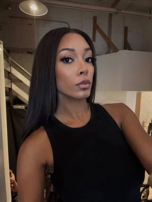 A woman with long, straight black hair taking a selfie in an indoor setting with lighting and wooden structures visible in the background.